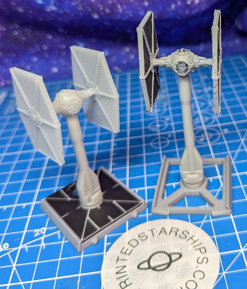 May include: Two gray TIE fighter models. One has two solar panel wings and the other has two black solar panel wings. Both models are on gray stands with a square base. A white circular logo with text is in the foreground.