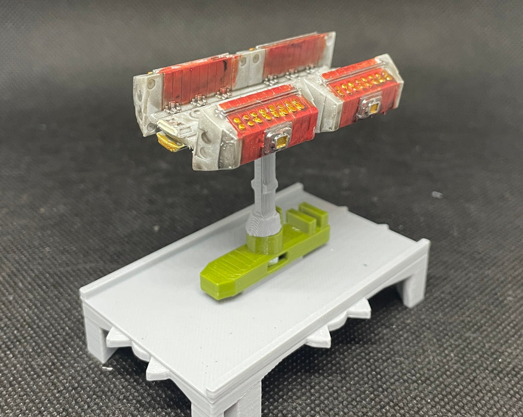 May include: A small, gray, plastic platform with a green, plastic base and a white and red, plastic model of a spaceship on top. The model has a series of small, gold details.
