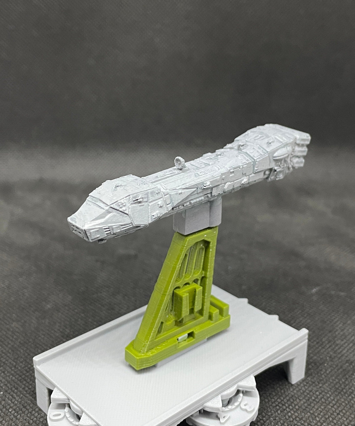 May include: A gray and green 3D printed model of a spaceship on a gray base. The spaceship is on a green stand with a rectangular base and a triangular top.