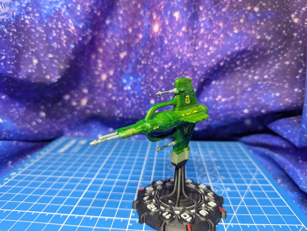 May include: A green spaceship model with a number 2 on top. The spaceship has a long, thin body with two side extensions and is mounted on a black and gray base. The background is a purple and blue galaxy pattern.