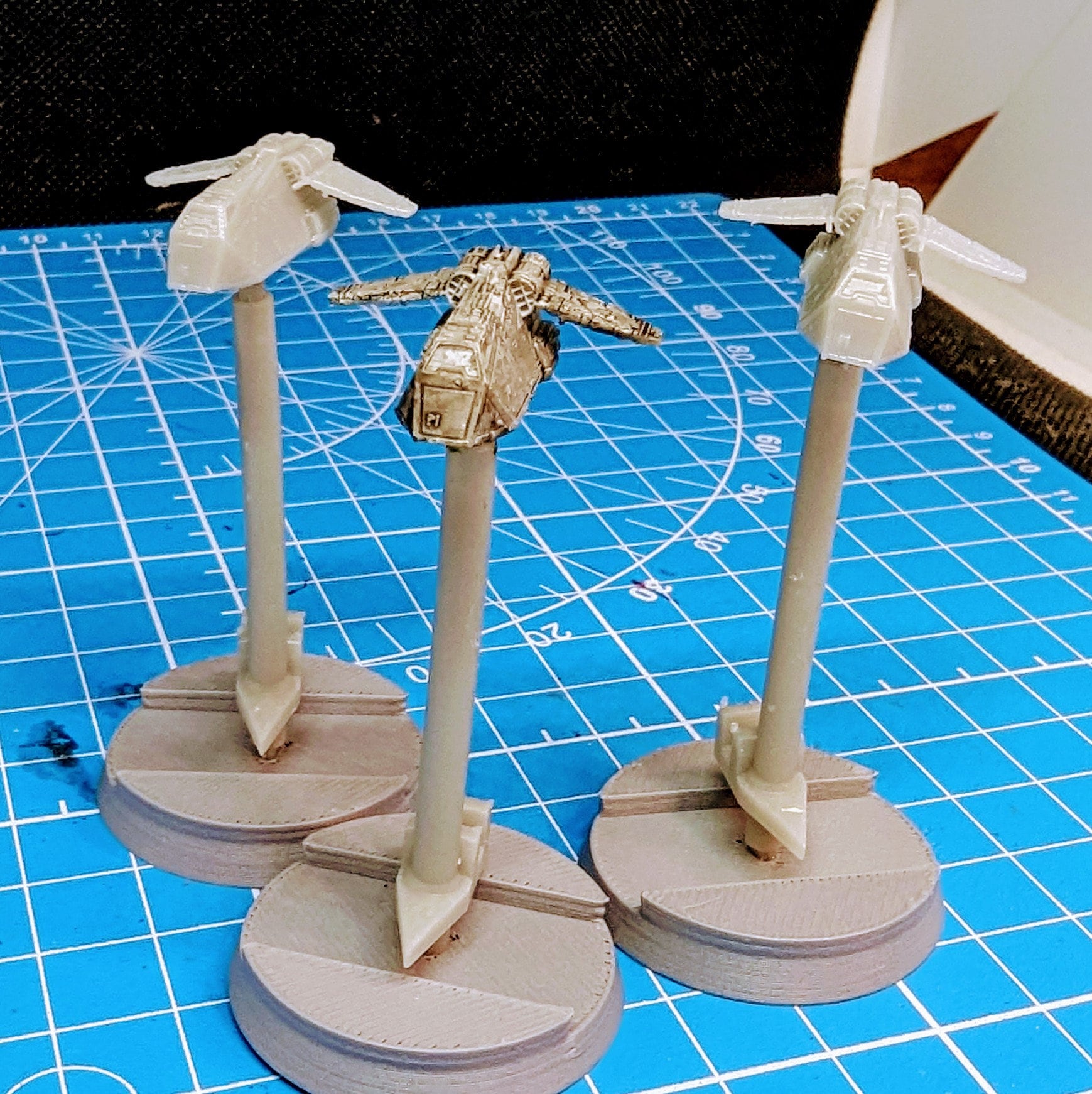 May include: Three miniature spaceships on gray bases with white stems. The spaceships are gray and have a rounded shape. The bases are circular and have a textured surface. The spaceships are arranged in a row on a blue surface with white grid lines.
