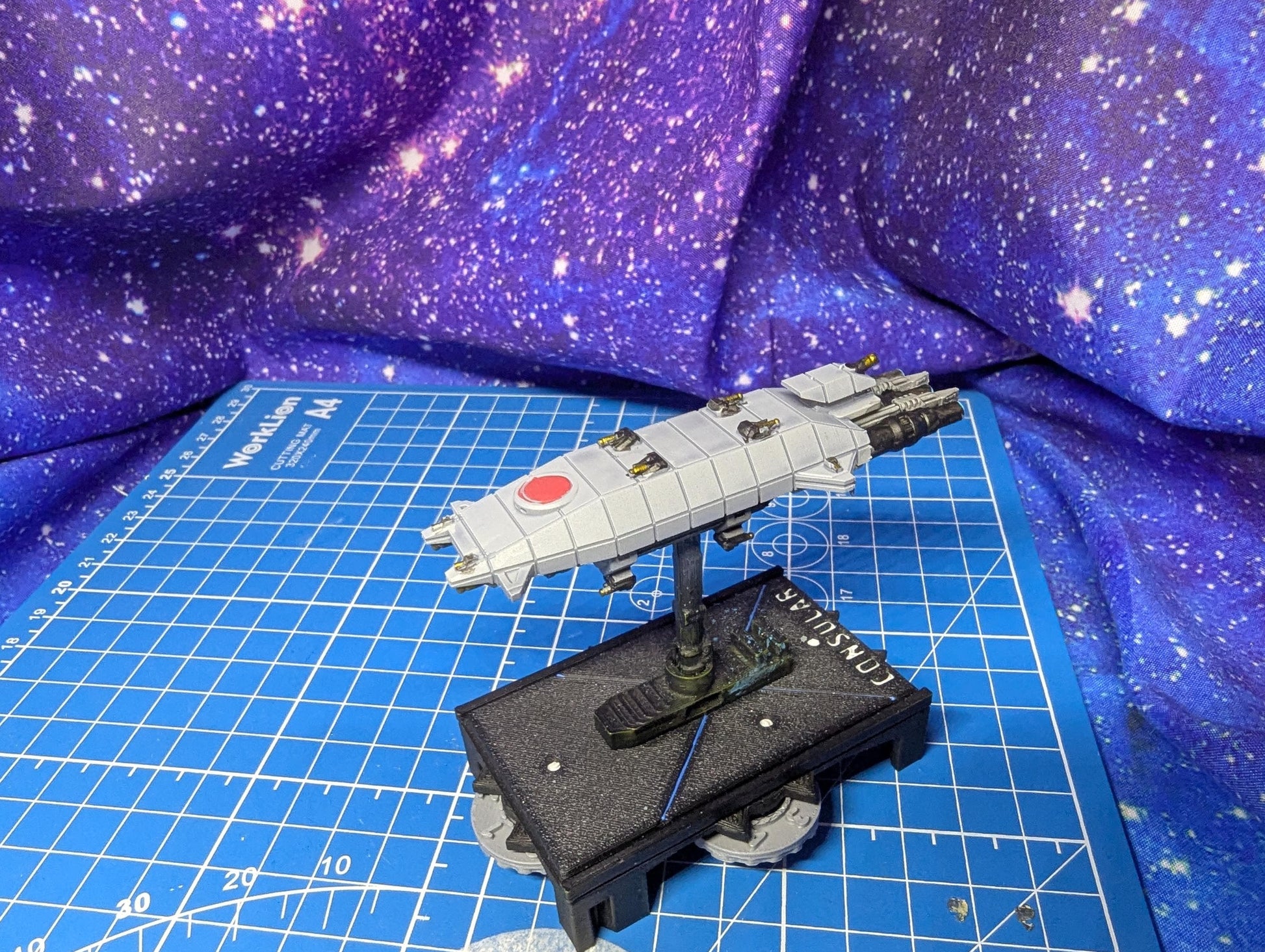 May include: A model of a Consular-class cruiser starship. The ship is primarily light gray with a red circle marking. It is mounted on a black base with the word 'Consular' printed on it. The background is a blue cutting mat and a purple galaxy fabric.