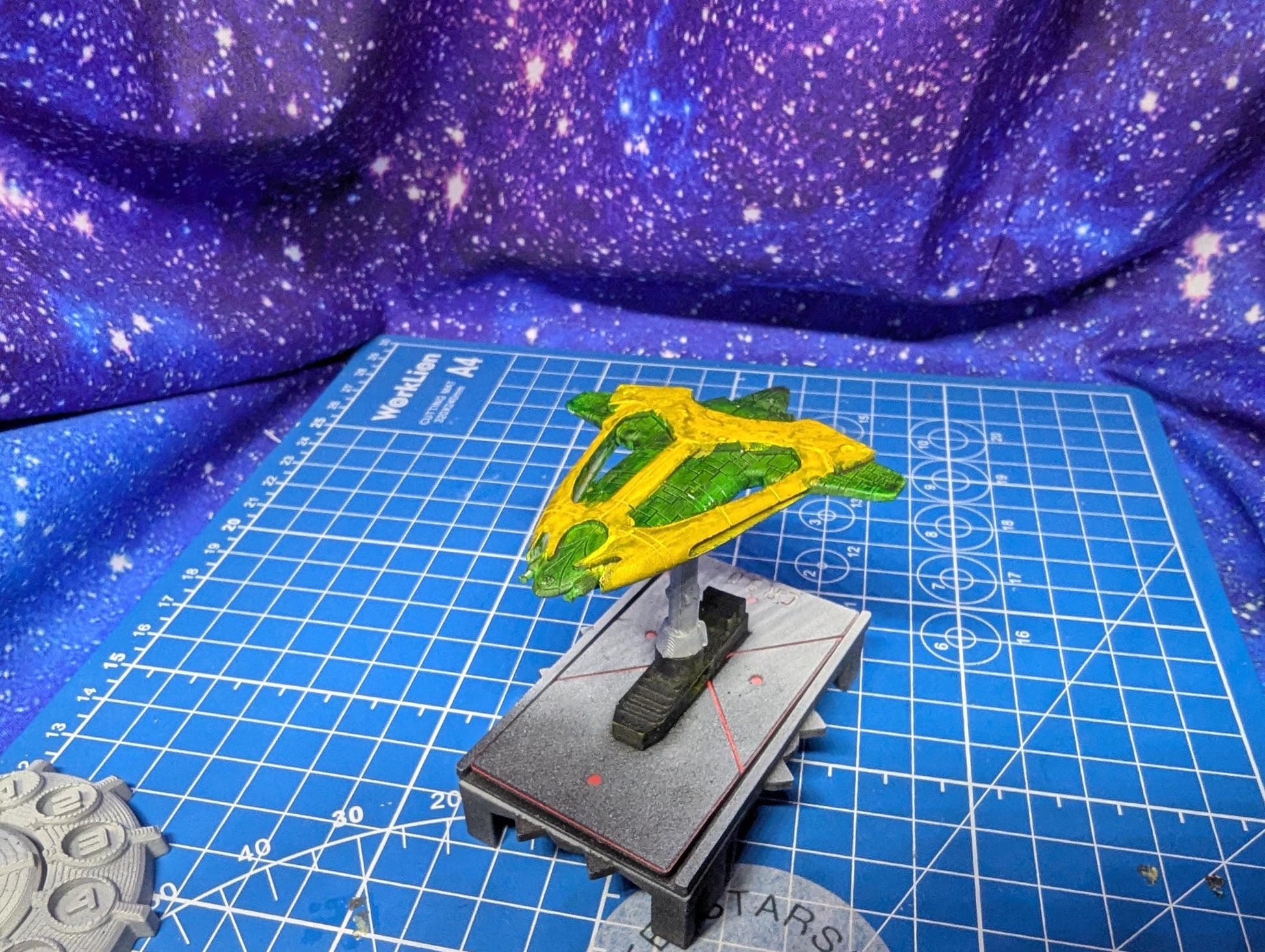 May include: A yellow and green spaceship model with a triangular design. The model is mounted on a gray and black base with a stand. The spaceship is set against a blue grid background with a purple and white galaxy design.