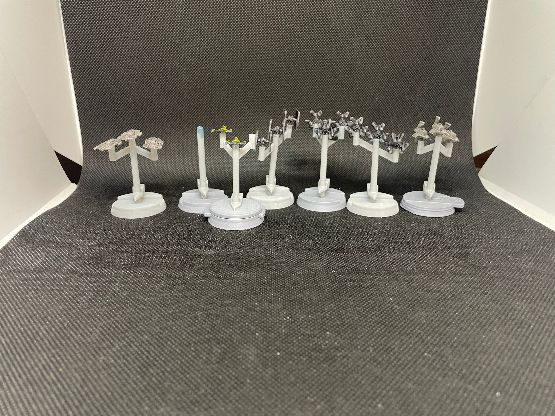 May include: A collection of nine miniature spaceships on stands. The spaceships are painted in shades of gray and black. The stands are circular and gray. The spaceships are arranged in a row on a black surface.