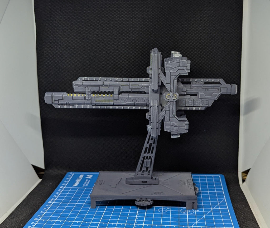 May include: A gray, detailed model of a spaceship with a long, rectangular body and a series of small, rectangular windows. The spaceship is mounted on a gray stand with a black base. The model is displayed on a blue and white grid surface.