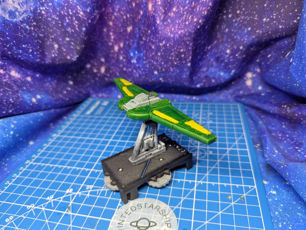 May include: A green and yellow spaceship model. The model has a wing-like design with yellow accents. It sits on a black base with silver supports, placed on a blue grid surface. The background features a purple and blue galaxy-like pattern.
