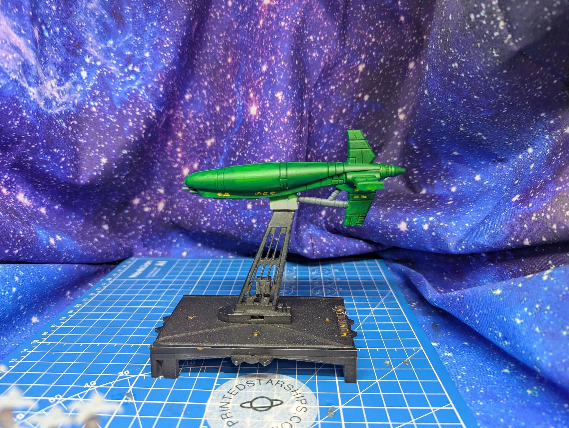 May include: A green spaceship model is displayed on a black stand against a blue background with white specks. The spaceship has a long, cylindrical body with a fin on top and wings. The stand is rectangular with a support structure. The background resembles a starry sky.