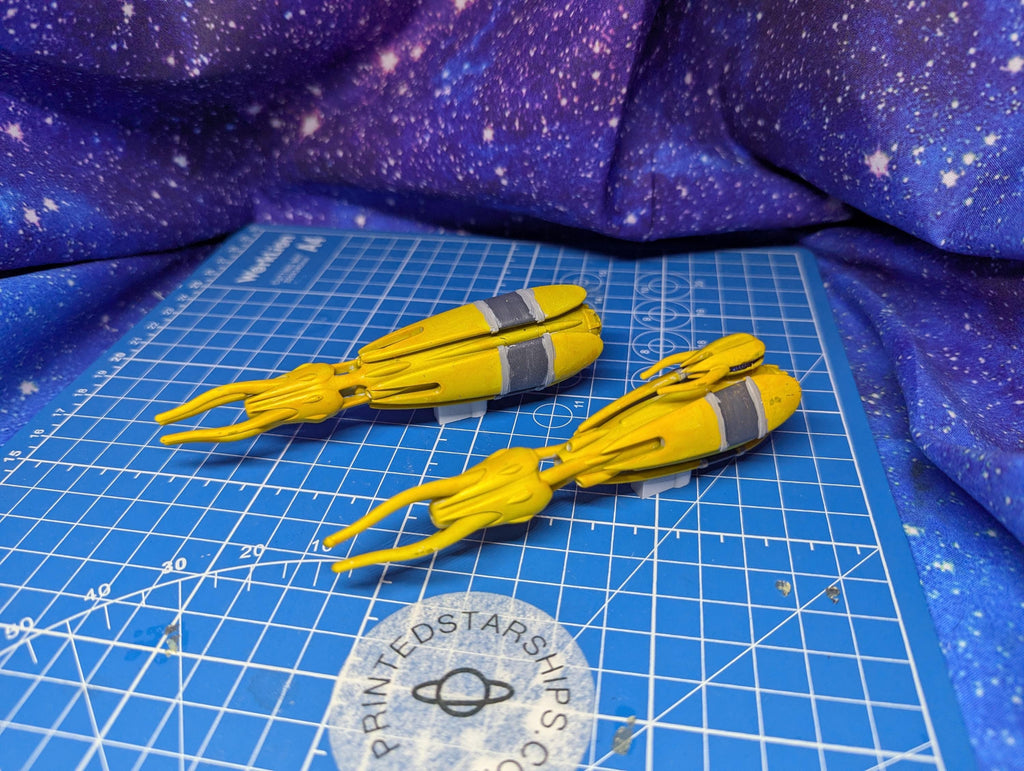 May include: Two yellow spaceship models with gray accents are displayed on a blue grid surface. The models have a streamlined design with elongated, tentacle-like appendages extending from the front. The background features a starry purple fabric and a logo that reads 'PRINTEDSTARSHIPS.COM'.