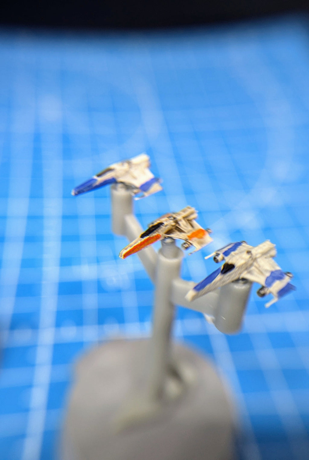 May include: Three miniature model spaceships on a gray stand. The spaceships are white, blue, and orange. The spaceships are on a blue grid background.