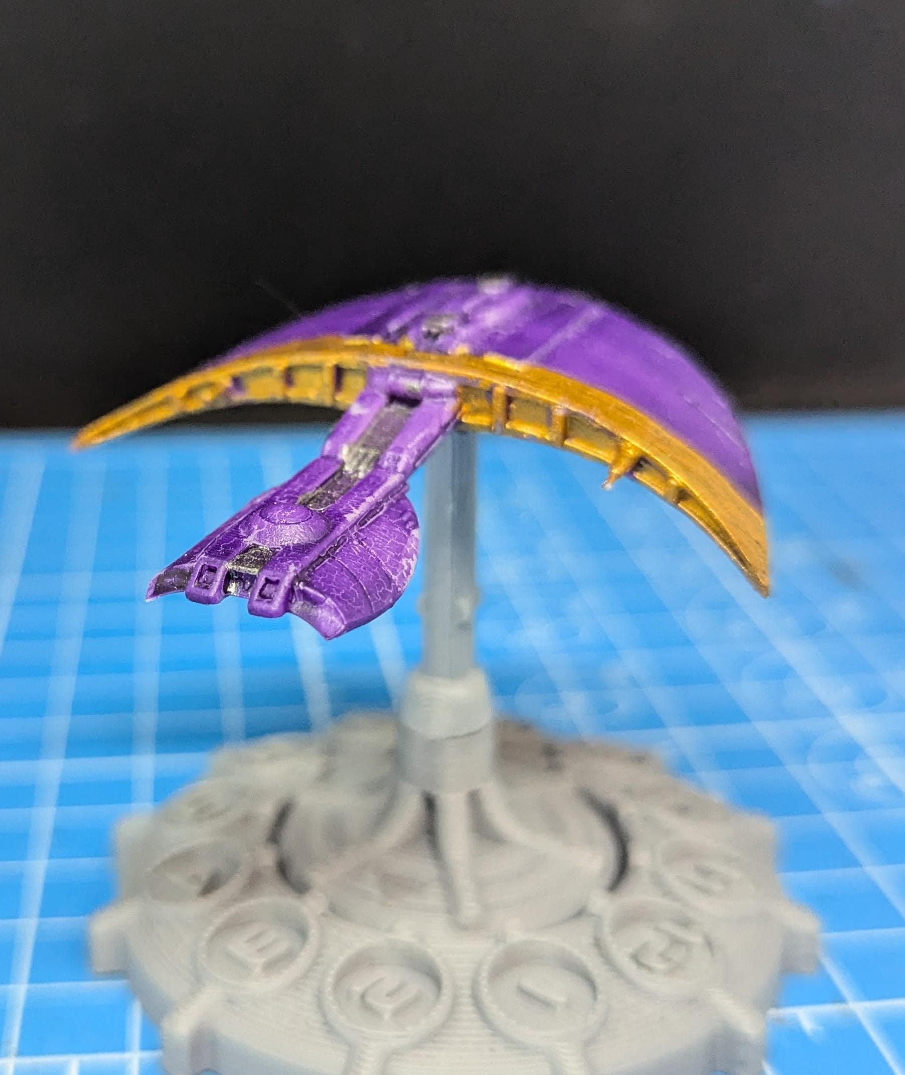 May include: A purple and gold spaceship model on a gray stand. The spaceship has a curved, wing-like design with a pointed nose. The wings are painted gold and the body is painted purple.