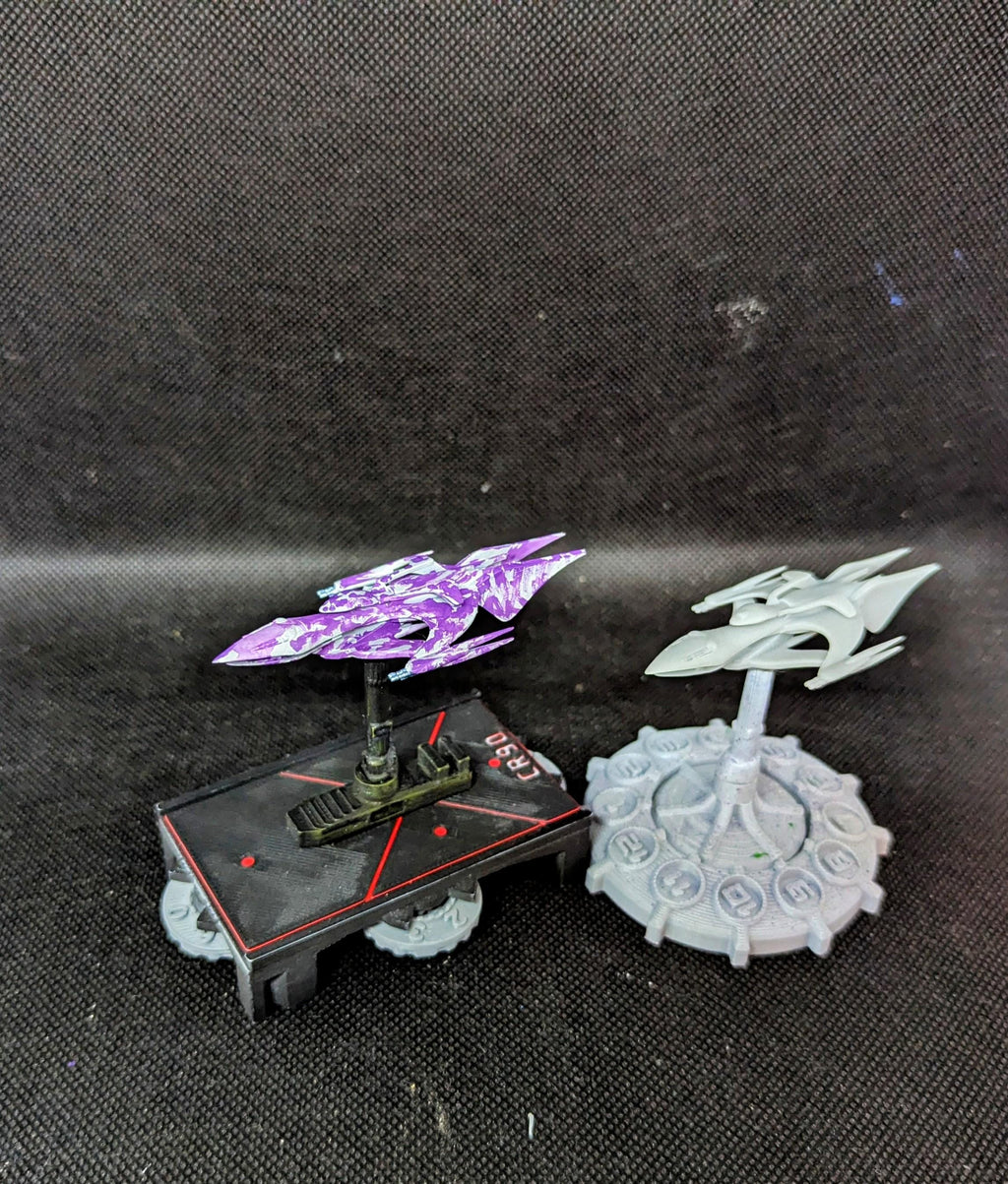 May include: Two miniature spaceships on bases. One spaceship is purple and white with a camouflage pattern. The other spaceship is white with a gray base. The base of the purple spaceship has the word 'CRSD' printed on it. The base of the white spaceship has numbers printed on it.