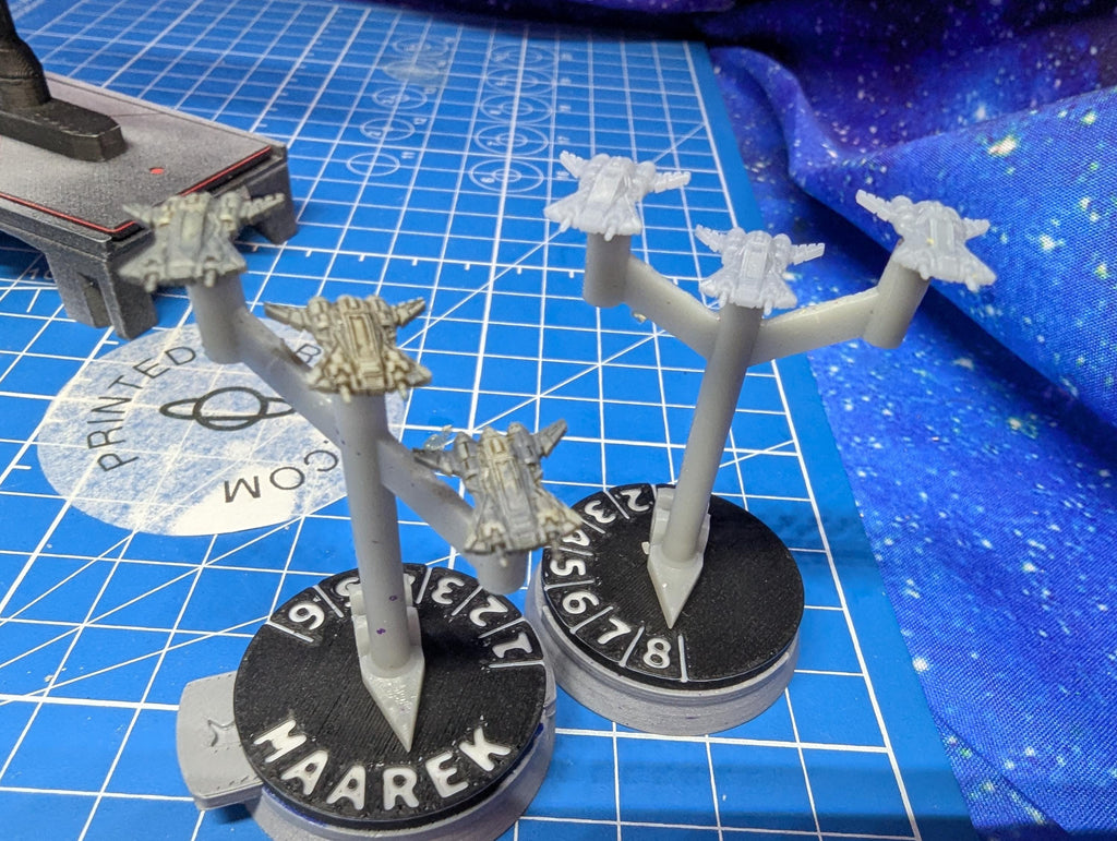 May include: Two miniature spaceship models are displayed on a blue grid surface. Each model is mounted on a black circular base with the word 'HAAREK' and numbers. The spaceships are gray with multiple wings and a central body. The bases have a white pointer.