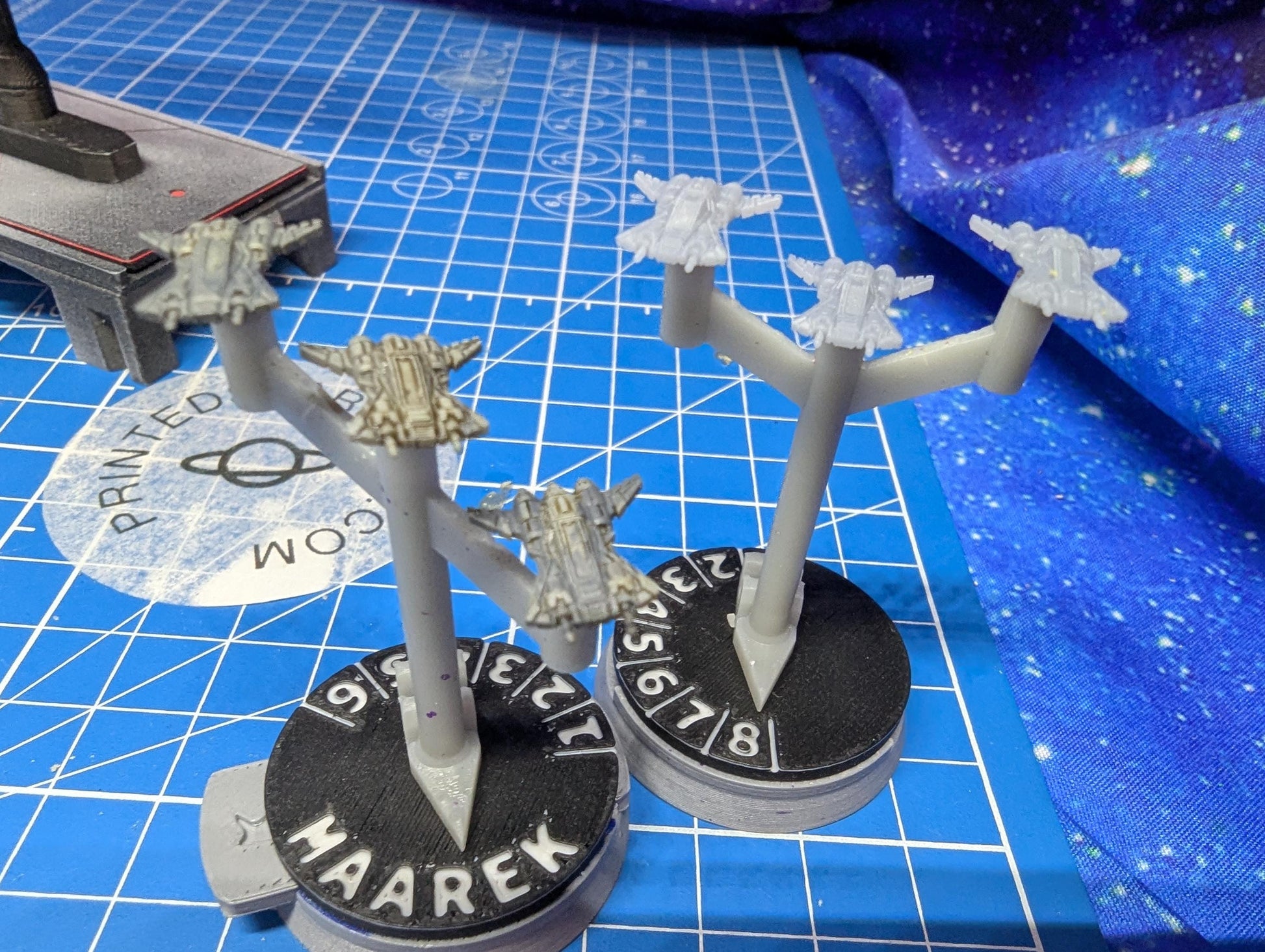 May include: Two miniature spaceship models are displayed on a blue grid surface. Each model is mounted on a black circular base with the word 'HAAREK' and numbers. The spaceships are gray with multiple wings and a central body. The bases have a white pointer.