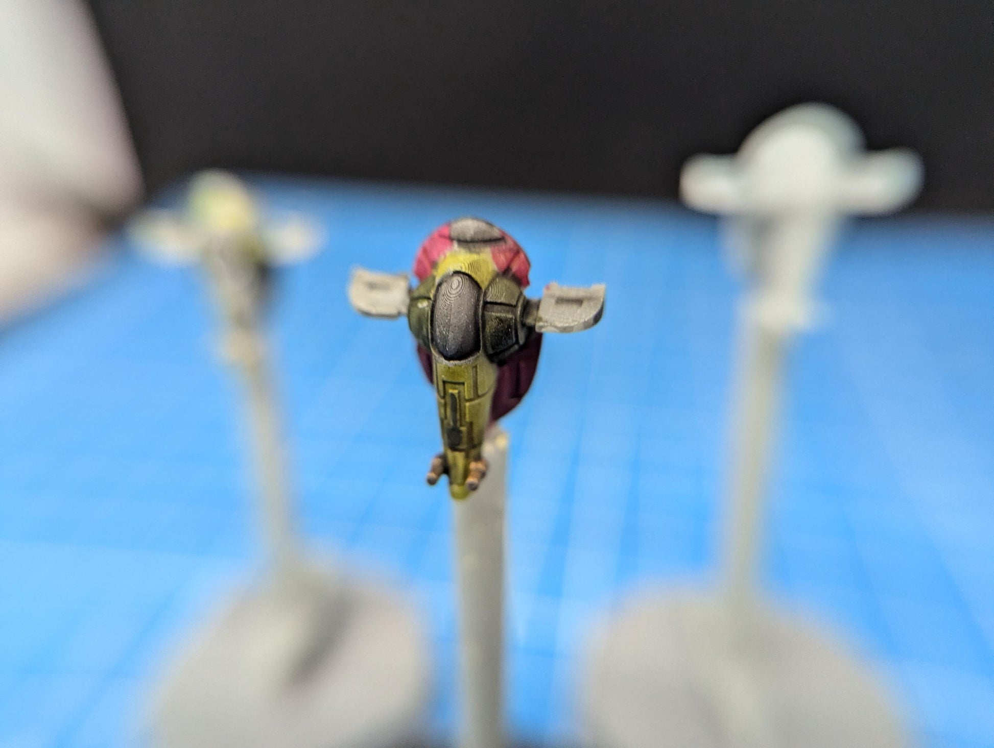 May include: A yellow and green miniature spaceship with a pink and red top. The spaceship has a clear cockpit window and is mounted on a white stand.