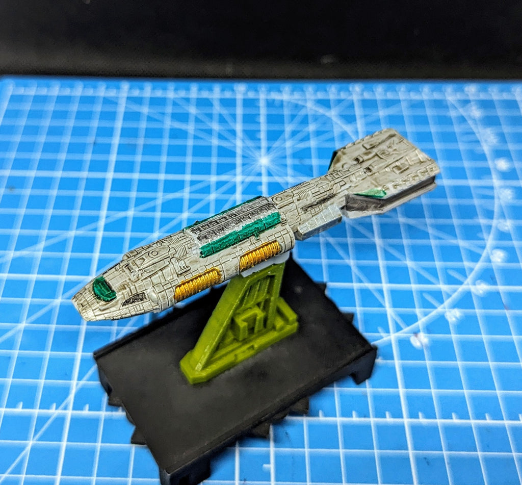 May include: A white and gray spaceship model with green and yellow details. The spaceship is mounted on a green stand and is displayed on a blue grid background.