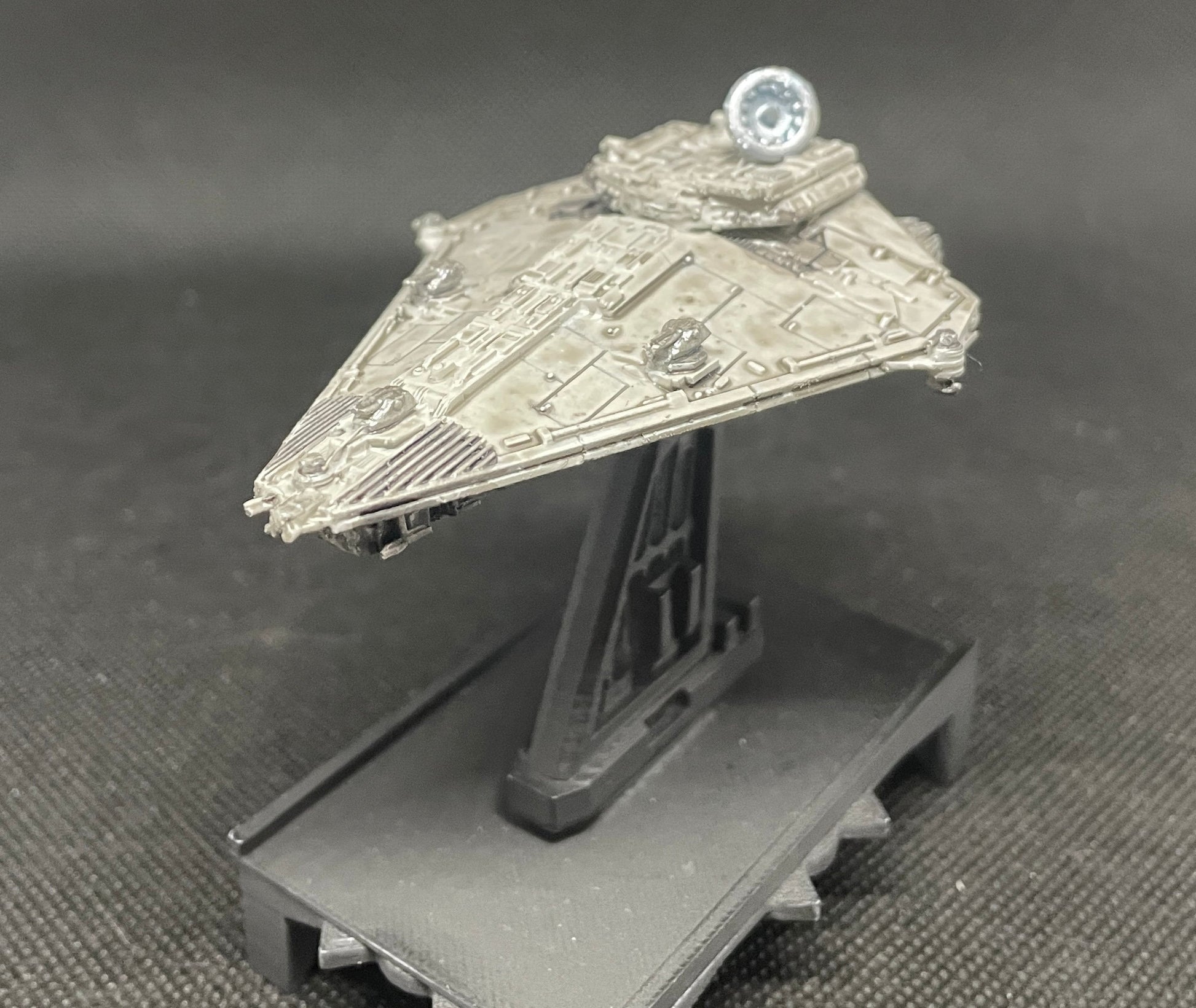 May include: A gray and silver spaceship model with a round, silver dome on top. The spaceship is on a black stand with a textured surface.
