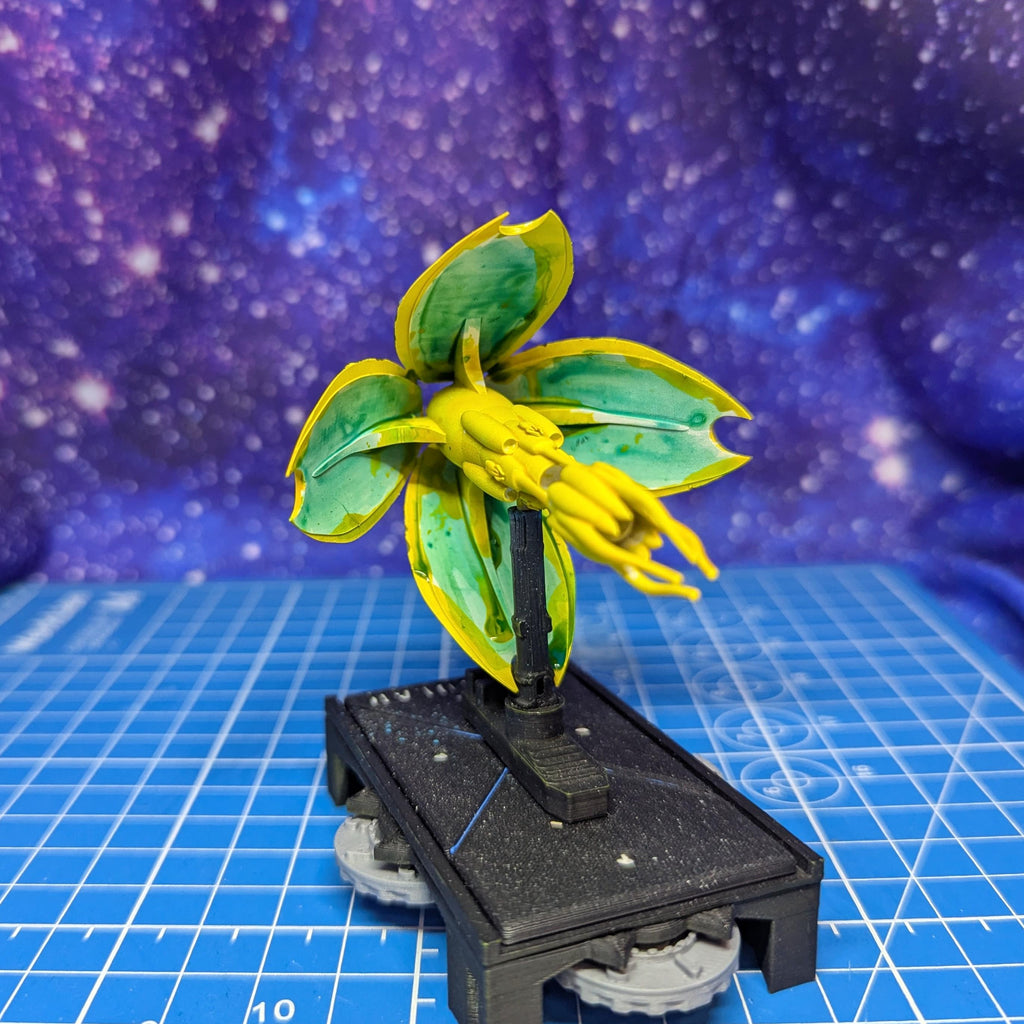 May include: A miniature model of a plant-like structure. The model features large, yellow and green leaves with a central yellow cluster. It is mounted on a black base with small, gray wheels, suggesting a tabletop game piece or decorative item. The background is a blue surface with a galaxy pattern.