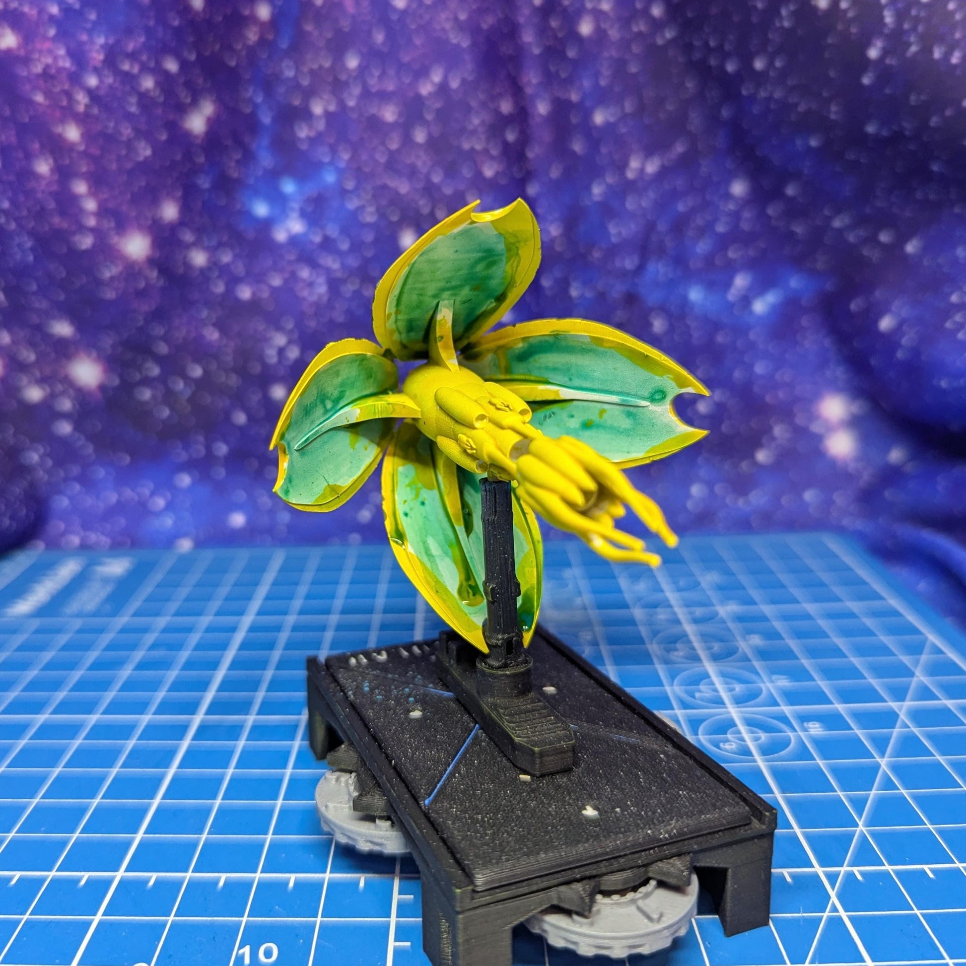 May include: A miniature model of a plant-like structure. The model features large, yellow and green leaves with a central yellow cluster. It is mounted on a black base with small, gray wheels, suggesting a tabletop game piece or decorative item. The background is a blue surface with a galaxy pattern.