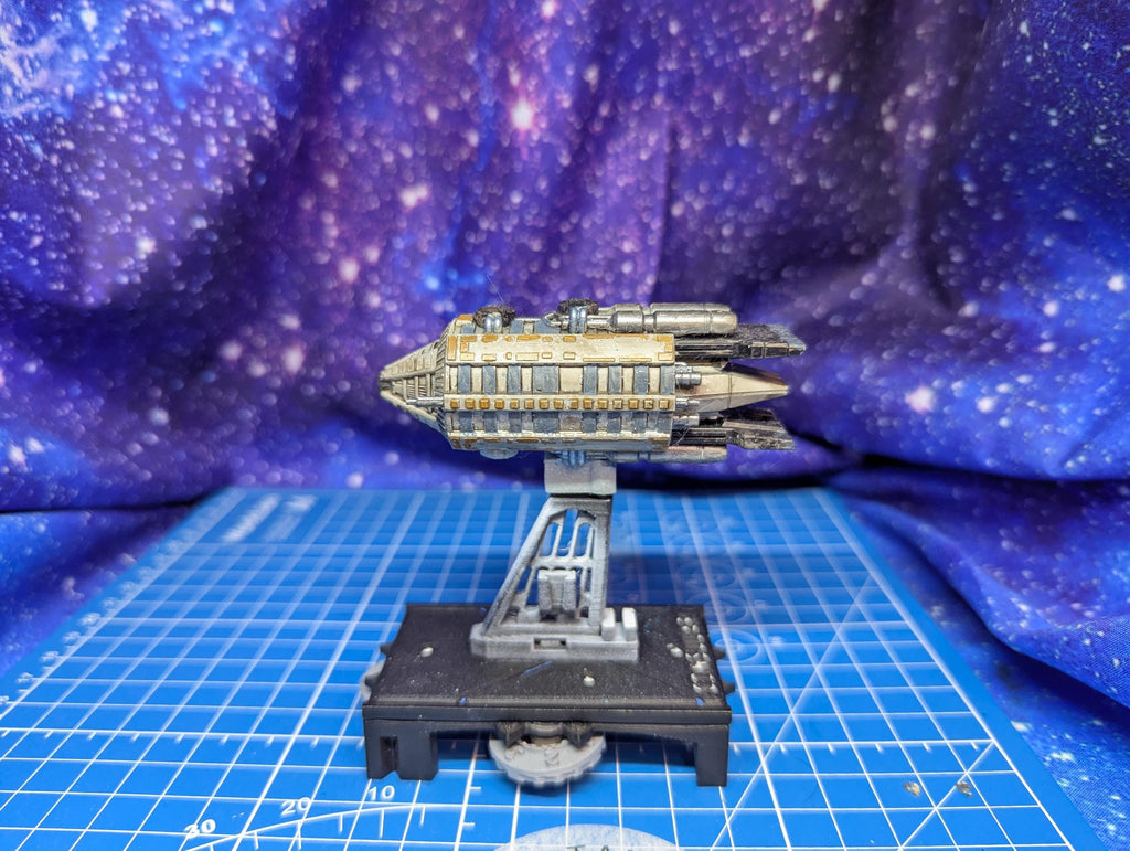 May include: A miniature spaceship model in shades of gray, beige, and silver, mounted on a gray stand with a black base. The spaceship features a rectangular body with detailed paneling and multiple engine-like protrusions. The background is a blurred purple and blue galaxy-themed fabric.