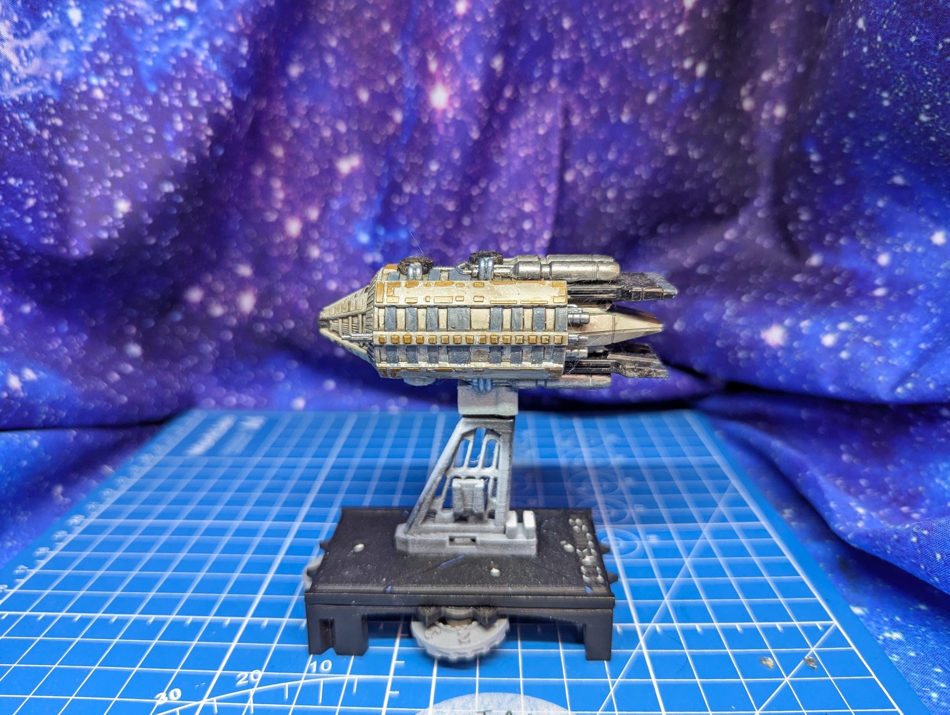 May include: A miniature spaceship model in shades of gray, beige, and silver, mounted on a gray stand with a black base. The spaceship features a rectangular body with detailed paneling and multiple engine-like protrusions. The background is a blurred purple and blue galaxy-themed fabric.