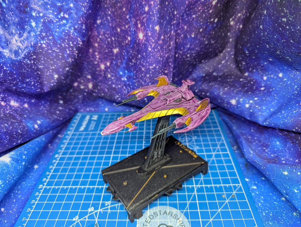 May include: A miniature starship model in shades of purple and gold, mounted on a black base. The ship features a sleek, angular design with prominent wings and a detailed surface. The base has gold accents and text. The background is a blue and purple galaxy-themed fabric.