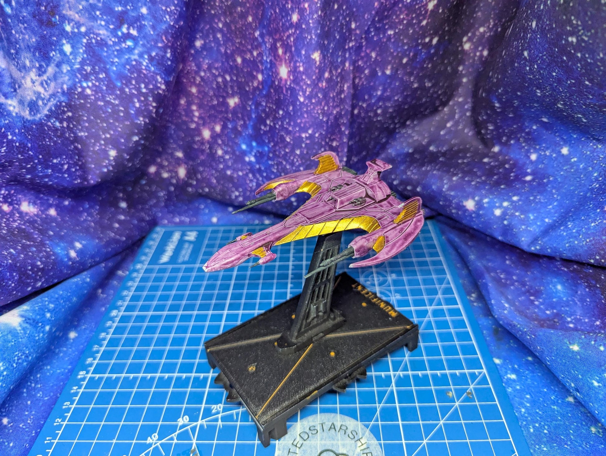 May include: A miniature starship model in shades of purple and gold, mounted on a black base. The ship features a sleek, angular design with prominent wings and a detailed surface. The base has gold accents and text. The background is a blue and purple galaxy-themed fabric.