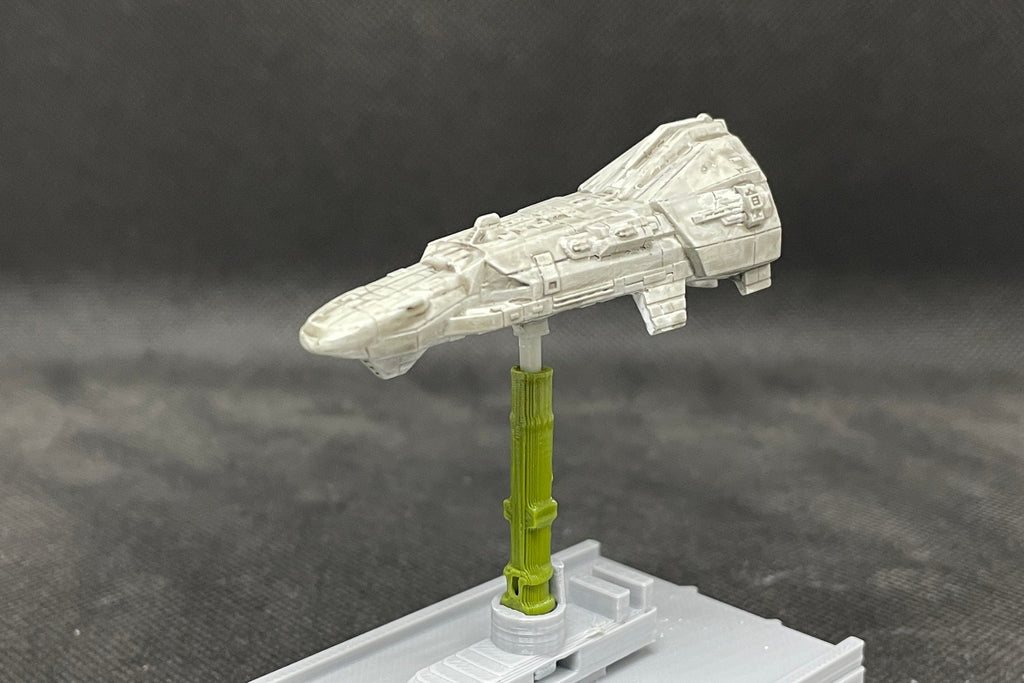 May include: Close-up view of a gray spaceship model, meticulously detailed with various textures and panels. It's mounted on a small, olive-green stand, which is attached to a gray base. The model's design suggests a science fiction or space-themed aesthetic, possibly from a miniature wargame or collectible series. The spaceship is angled slightly upward, showcasing its length and features.
