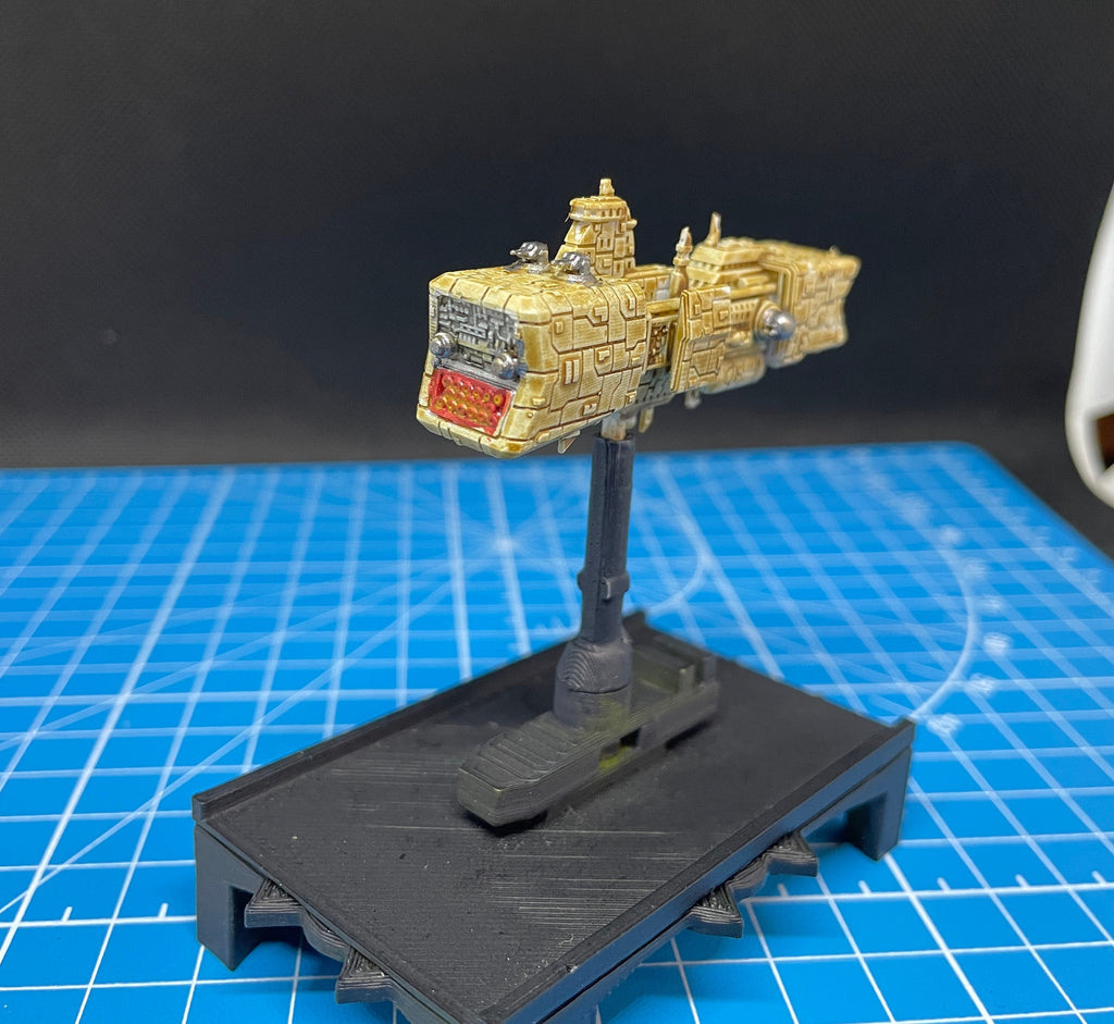 May include: A beige and brown spaceship model with a red light on the front. The spaceship is mounted on a black stand with a black base.