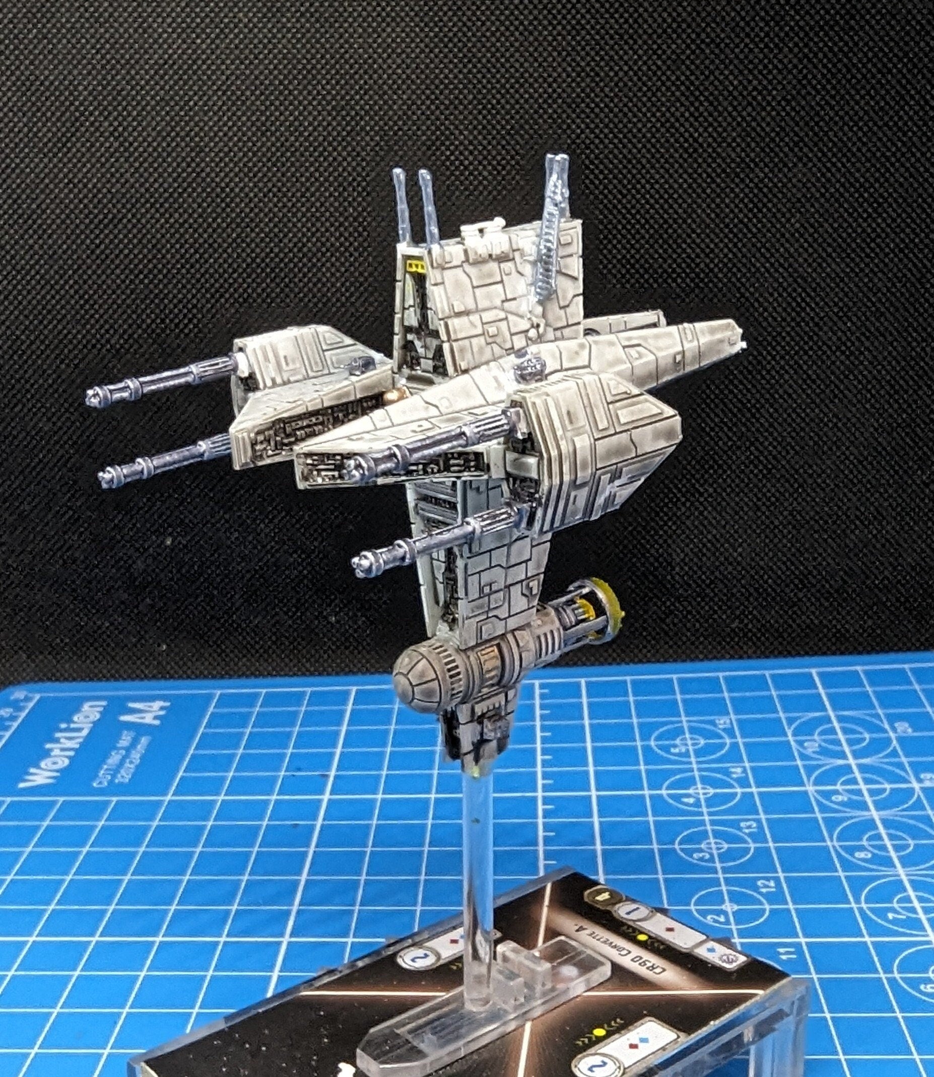 May include: A white and gray spaceship model with yellow accents. The spaceship has two long cannons on each side and a smaller cannon on the bottom. The spaceship is on a clear plastic stand and is on a blue and black gaming mat. The mat has numbers and circles printed on it. The spaceship is a Star Wars themed model.