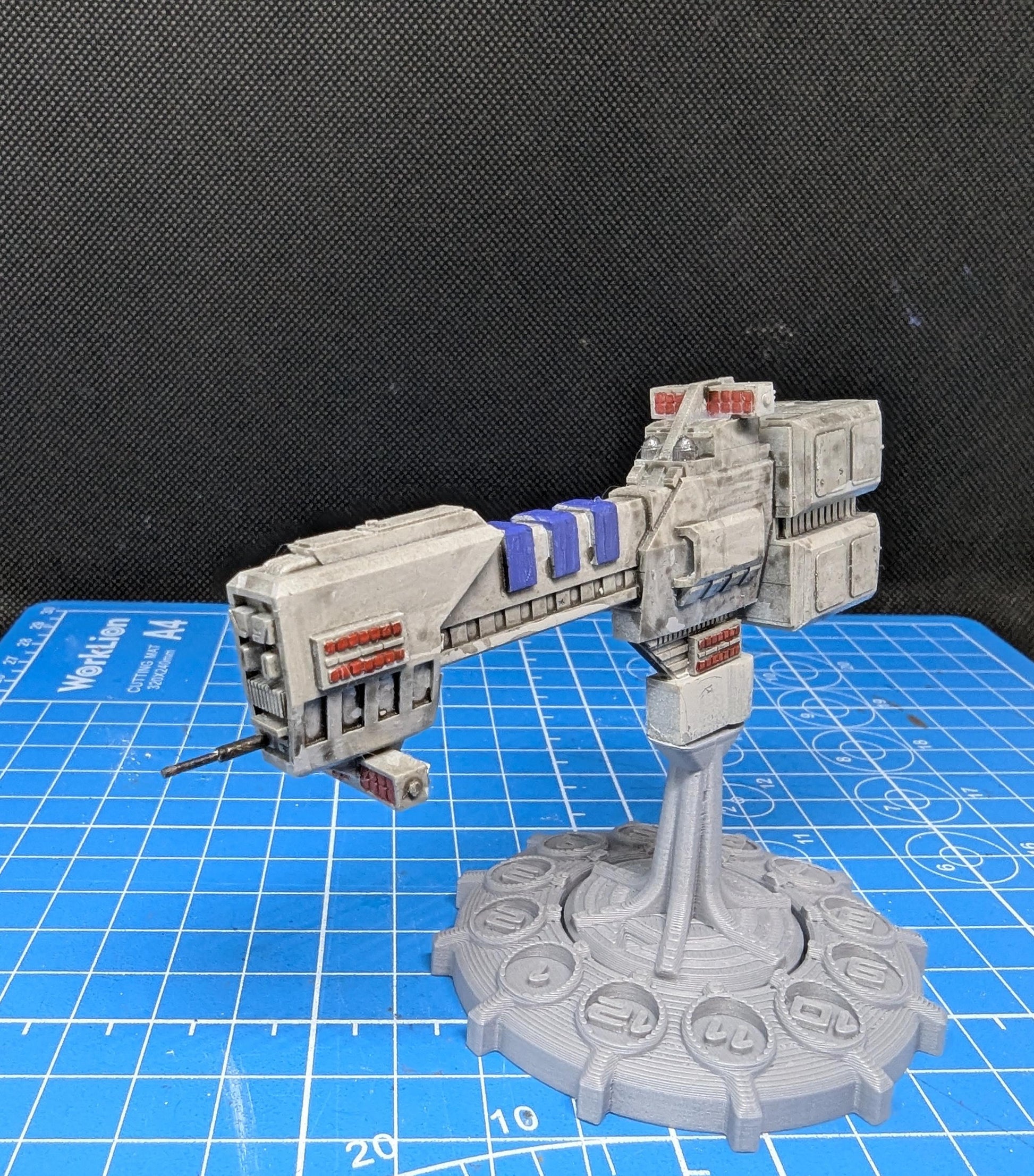 May include: Close-up view of a meticulously painted miniature spaceship model, predominantly white with accents of blue and red. The spaceship features detailed panel lines and various textures, suggesting a weathered or battle-worn aesthetic. It's mounted on a gray circular base with intricate detailing, possibly representing a launchpad or docking station. The model is set against a blue gridded surface, commonly used for crafting or miniature painting.