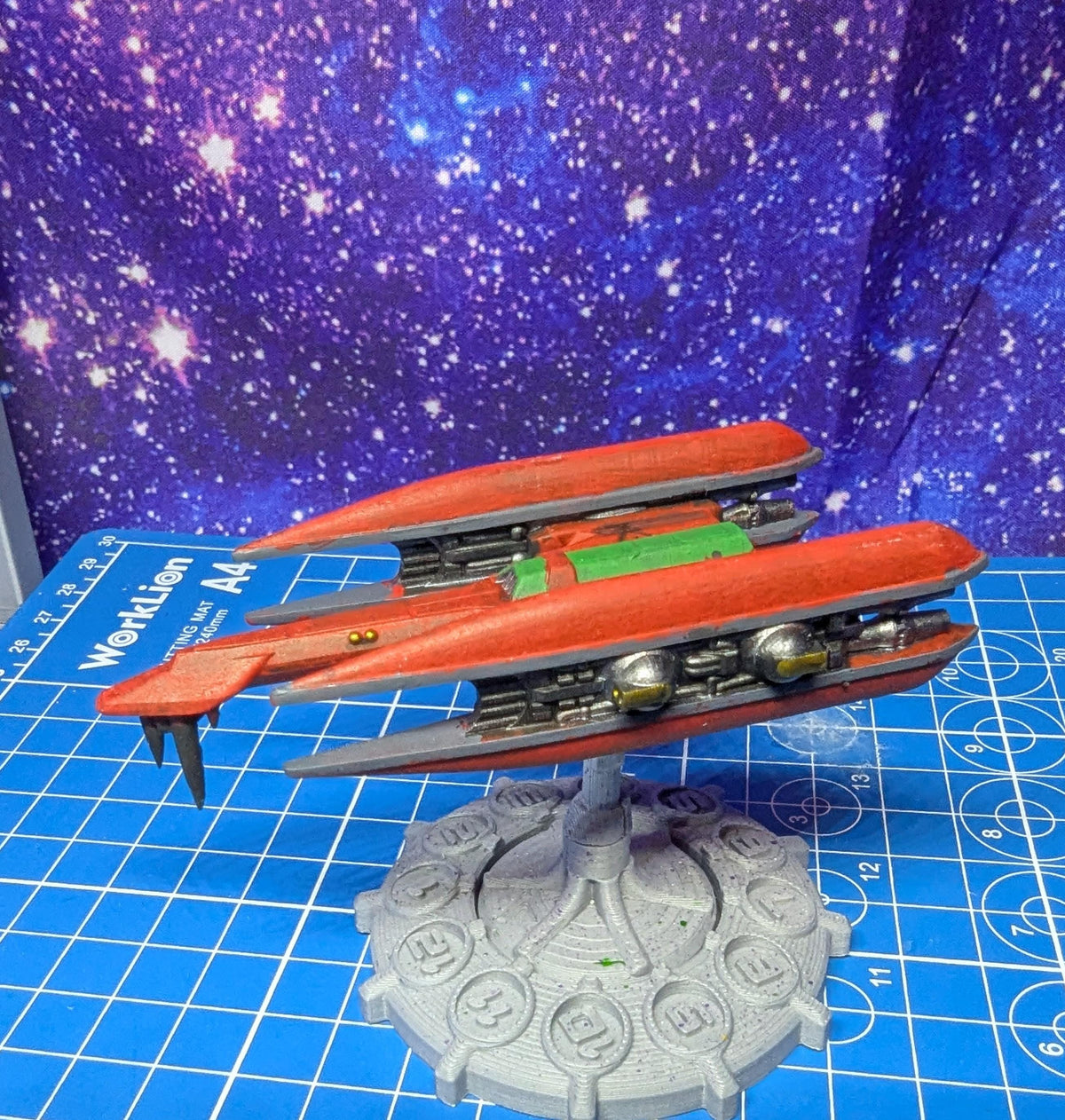 May include: A red and gray spaceship model with green details. The spaceship is on a gray base with a circular design and numbers 11 through 15. The spaceship is on a blue and white grid surface. The text 'WorkLion A4' is visible on the surface.