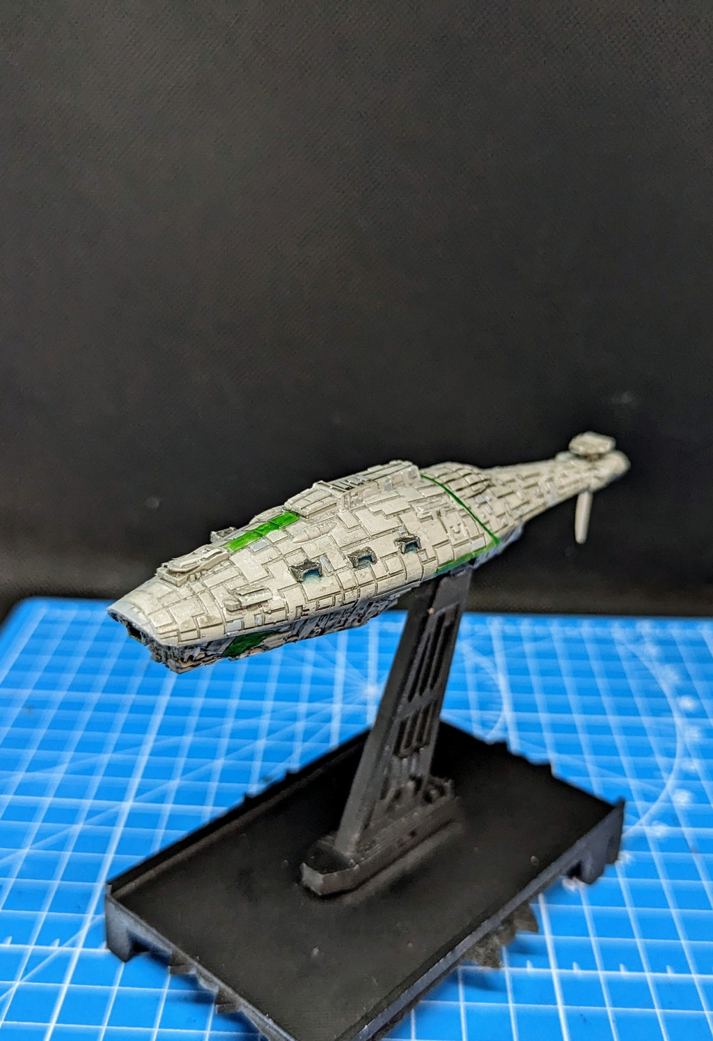 May include: A white and green spaceship model on a black stand. The spaceship has a long, narrow body with a pointed nose and a small, rectangular cockpit. The spaceship is painted with a light gray base coat and green accents. The stand is a simple, black plastic stand with a rectangular base and a single, vertical support.