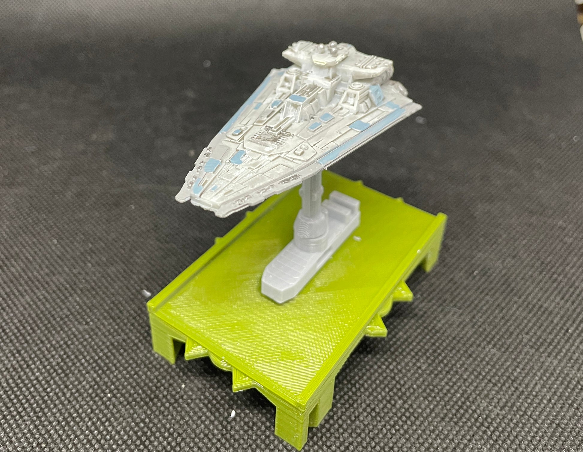 May include: A white and gray model of a starfighter spaceship on a green platform. The spaceship has blue accents and is positioned on a stand with a green base.