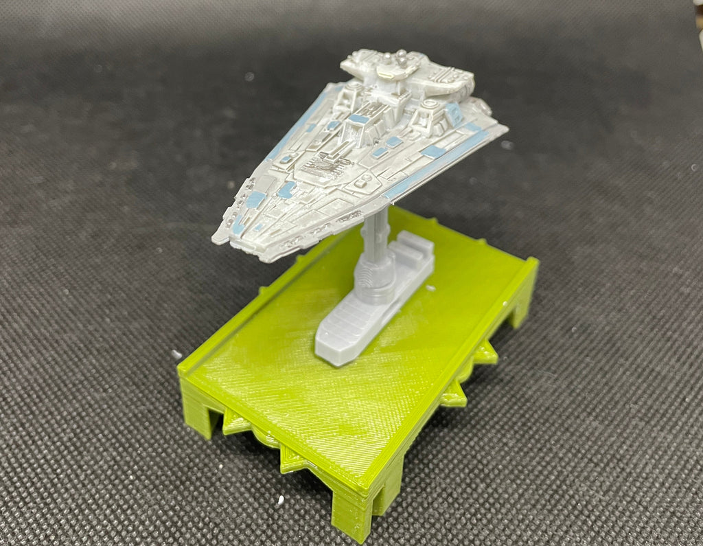 May include: A white and gray model of a starfighter spaceship on a green platform. The spaceship has blue accents and is positioned on a stand with a green base.