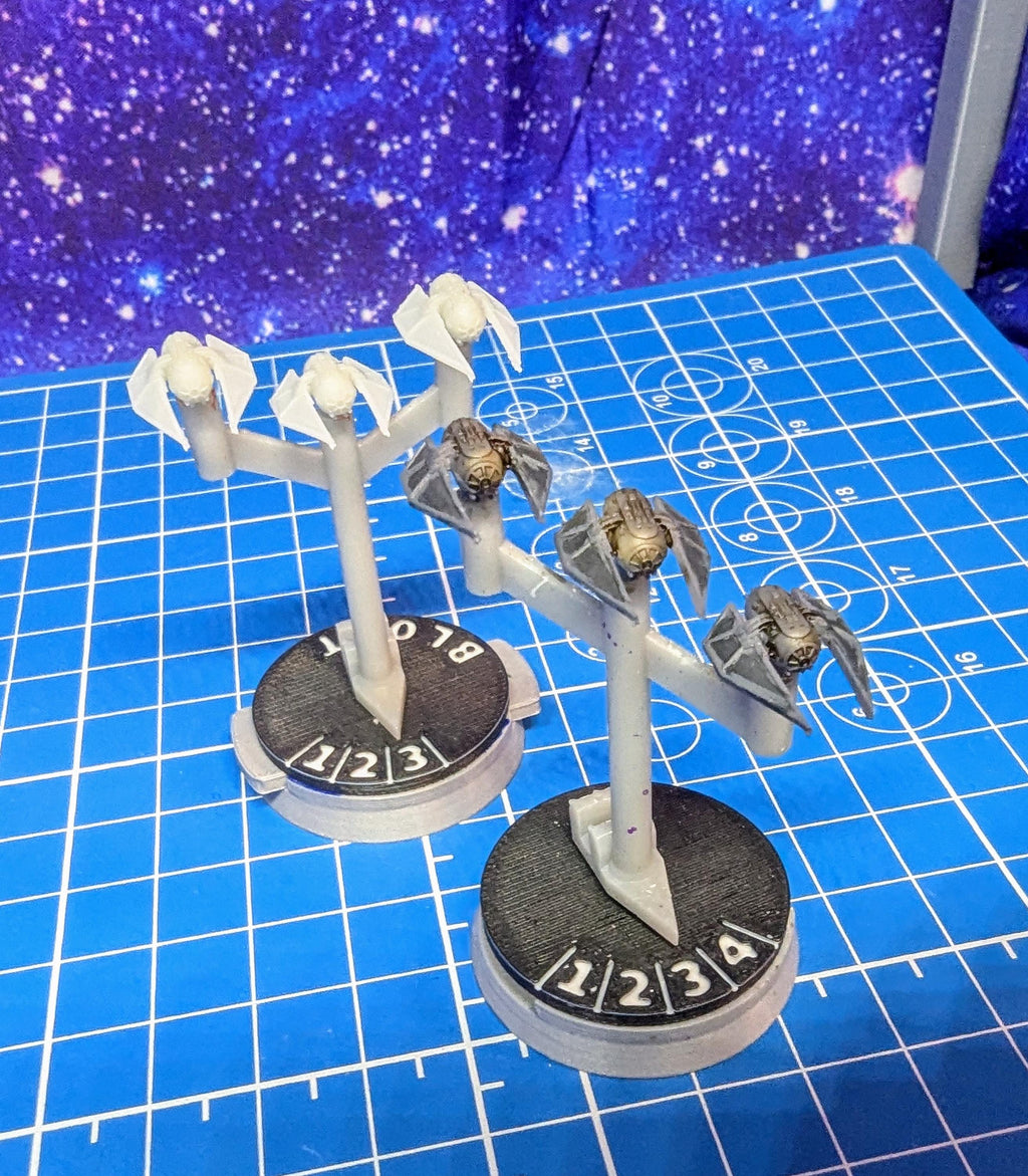 May include: Two sets of miniature spaceships on stands with black and white bases. The bases have numbers 1, 2, 3, and 4 printed on them. The spaceships are gray and have white wings. The spaceships are on a blue grid background.