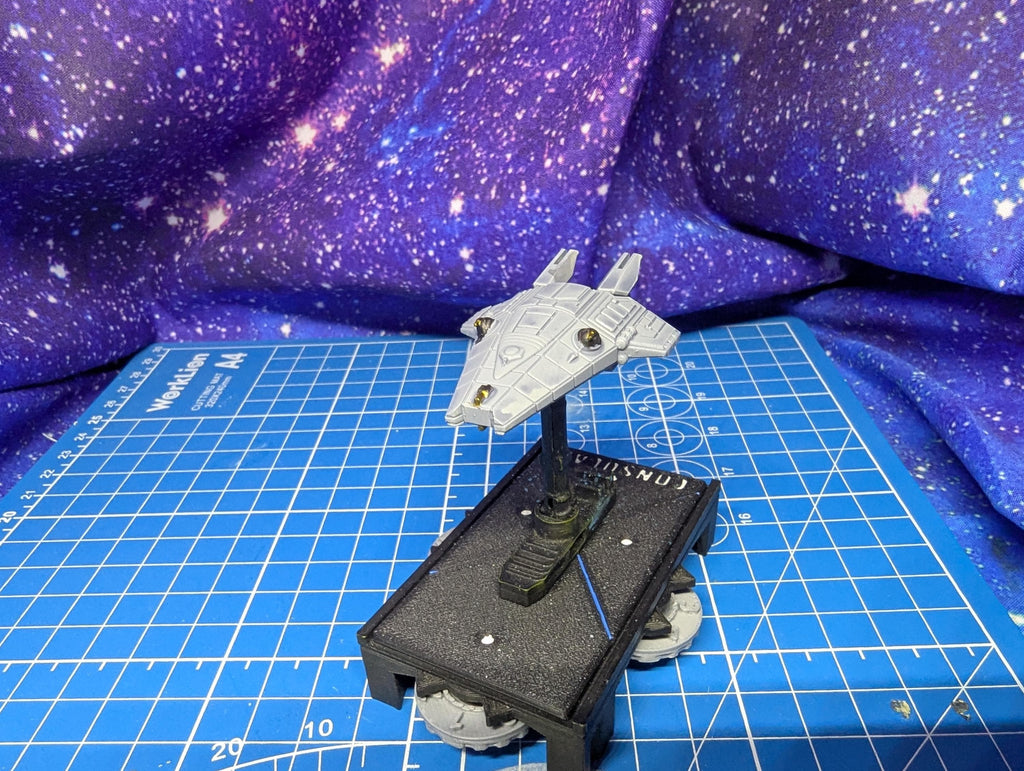 May include: A gray spaceship model with a rectangular base. The spaceship has a triangular body with two wings and is mounted on a black stand. The base is black with the word 'CONSULA' printed on it. The model is on a blue grid surface with a purple galaxy backdrop.