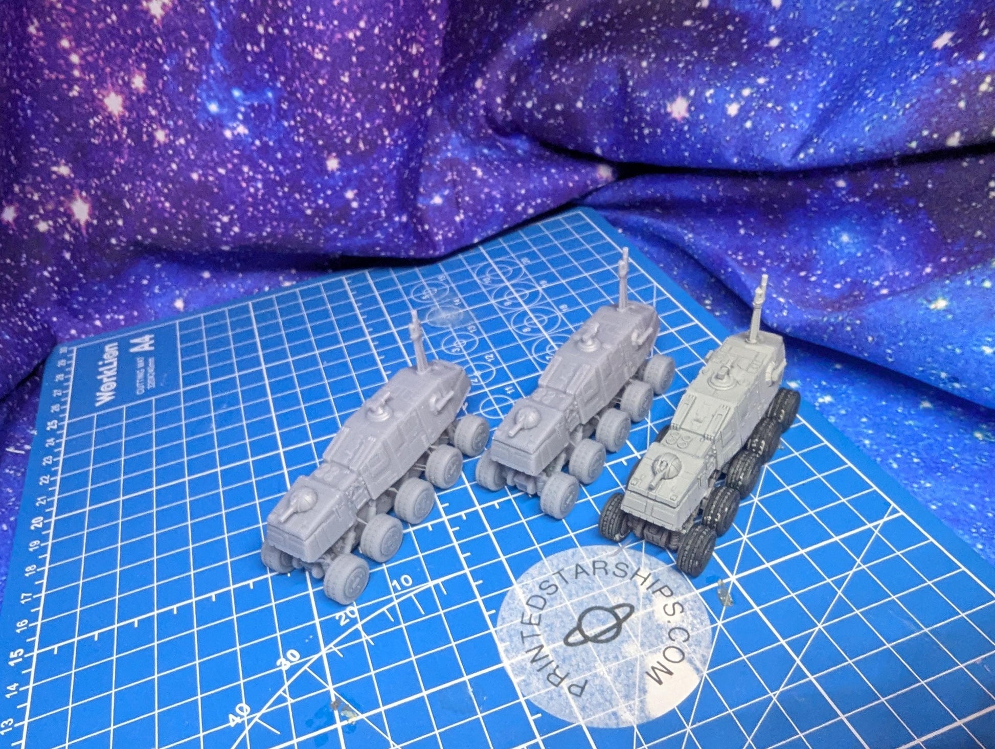 May include: Three gray, miniature, space rover models are displayed on a blue grid cutting mat. Each rover has a rectangular body, multiple wheels, and a tall antenna. The rovers are likely 3D printed and are a light gray color. A sticker with a planet and the text 'PRINTEDSTARSHIPS.COM' is visible.