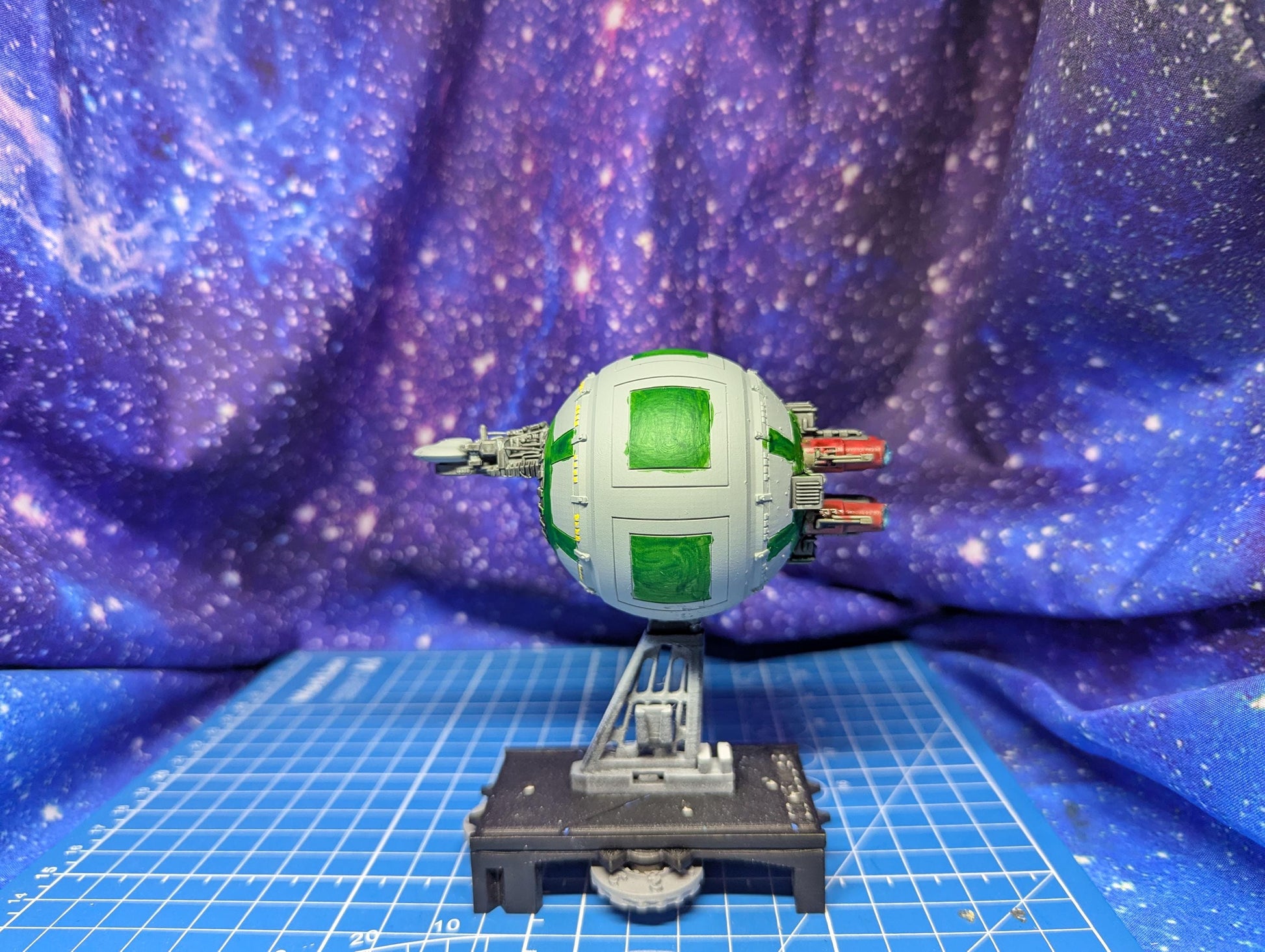 May include: A model spaceship with a spherical body in light gray and green. The sphere has green rectangular panels. It has two red engine pods and a small, gray, wing-like structure. The ship is mounted on a gray stand with a black base, set against a galaxy backdrop.