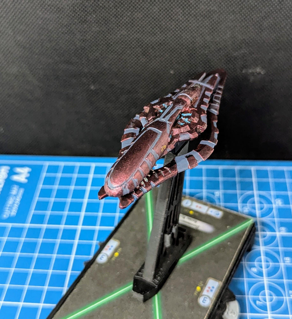 May include: A small, detailed, red and gray spaceship model with a black base. The spaceship has a long, pointed nose and a series of fins and wings along its sides. The model is painted with a metallic red paint and has gray details. The base is a simple black stand with a green laser beam pointing up from the base.