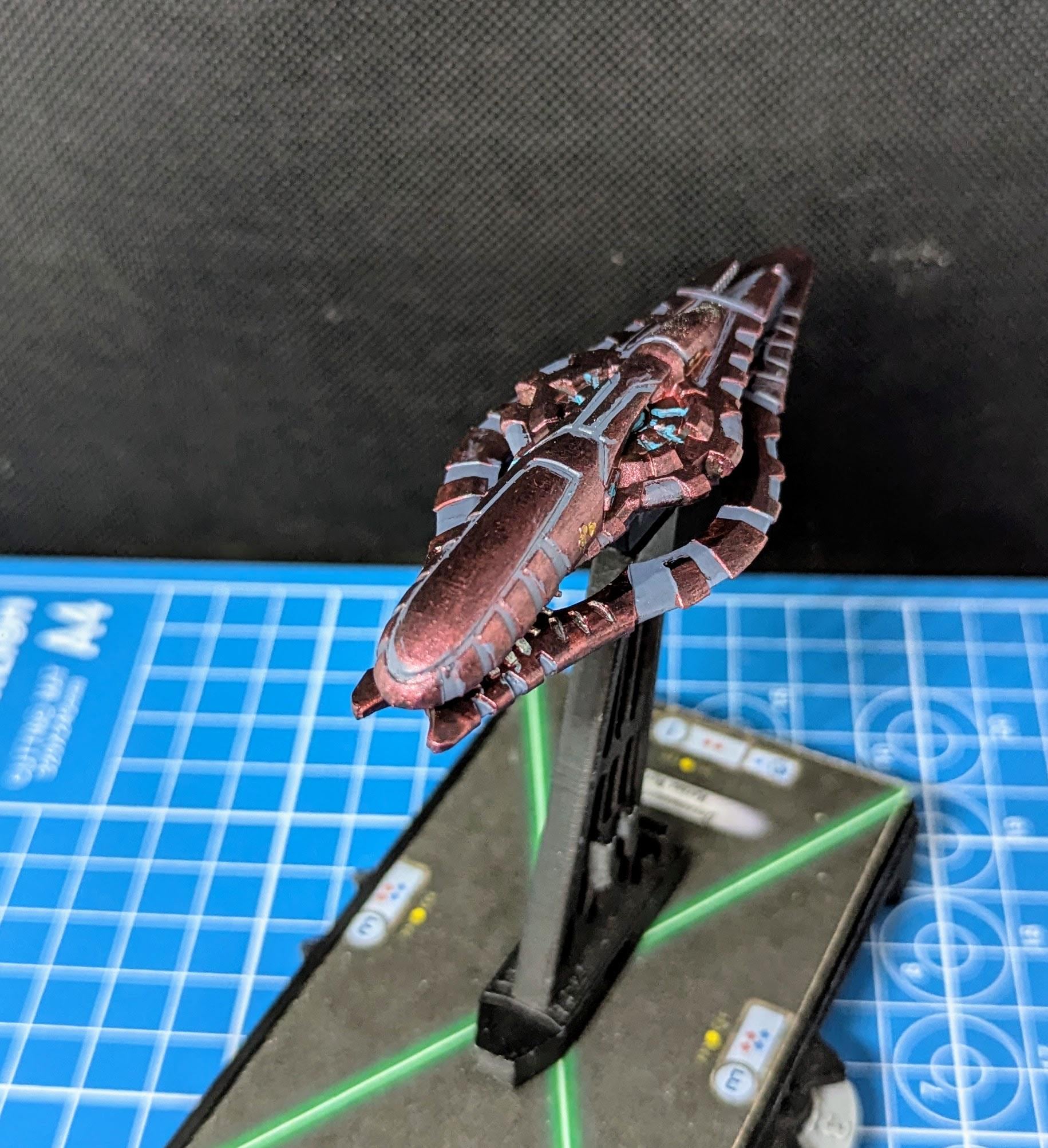 May include: A small, detailed, red and gray spaceship model with a black base. The spaceship has a long, pointed nose and a series of fins and wings along its sides. The model is painted with a metallic red paint and has gray details. The base is a simple black stand with a green laser beam pointing up from the base.