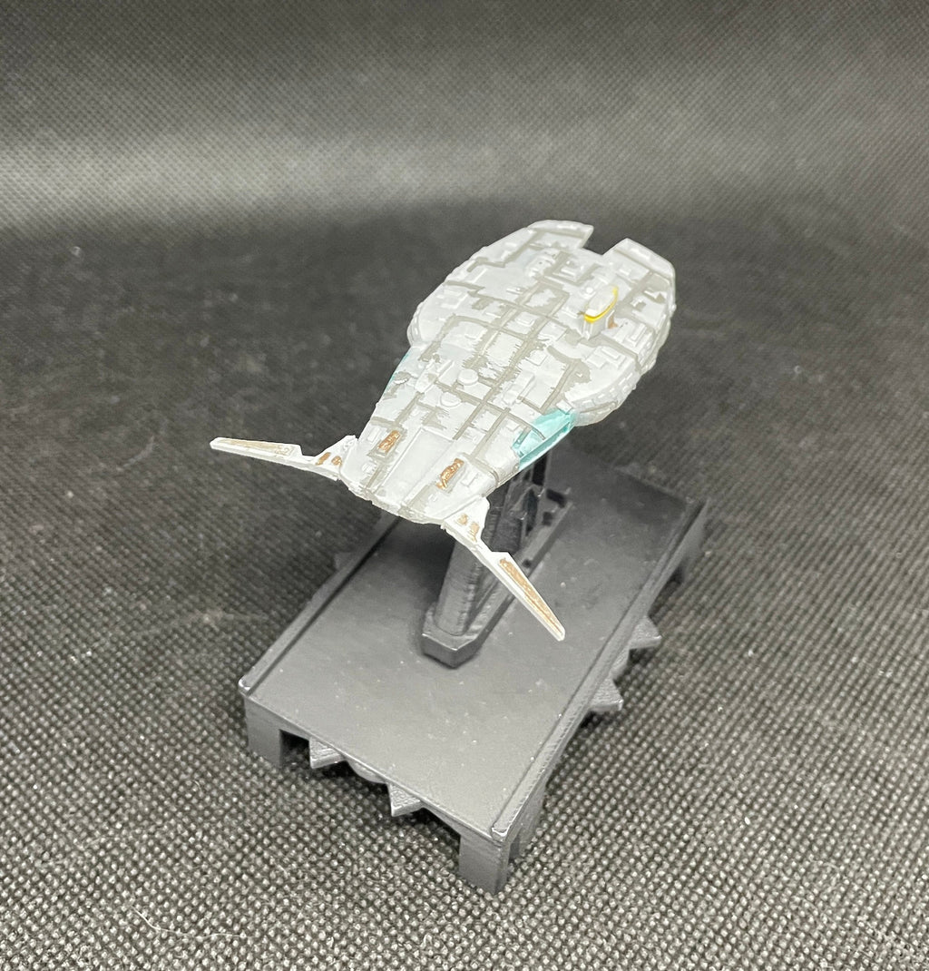 May include: A gray and white spaceship model with a teal and yellow accent. The spaceship is on a black stand with a textured surface.