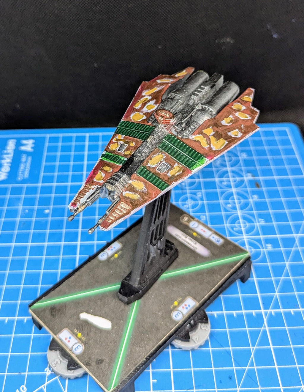 May include: Close-up view of a painted miniature starship model, predominantly brown and orange with green accents, mounted on a black display stand. The model features intricate details and a camouflage-like pattern.  The base includes markings and numbers. The miniature is displayed on a blue gridded surface.