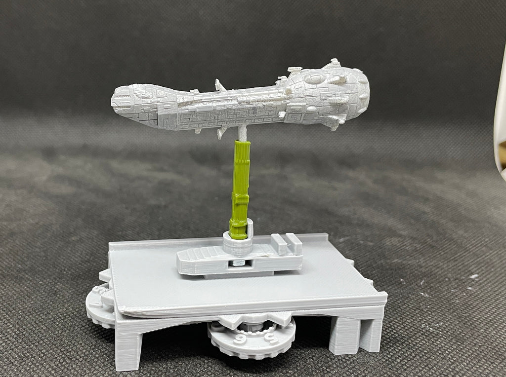 May include: A gray 3D printed model of a spaceship on a gray 3D printed base with a green support rod. The spaceship is long and narrow with a rounded front and a pointed back. The base has a circular platform with a gear on the bottom. The support rod is attached to the base and the spaceship.