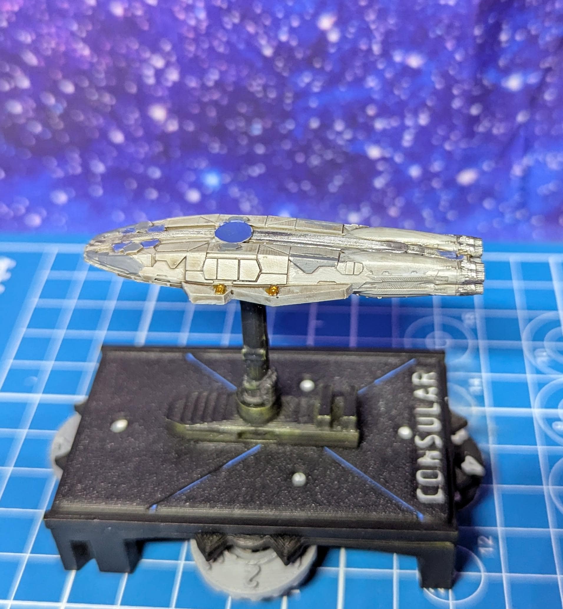 May include: A white and gray spaceship model with a blue circular detail on the top. The spaceship is mounted on a black base with the word 'CONSULAR' printed on the side. The base is on a blue grid surface.