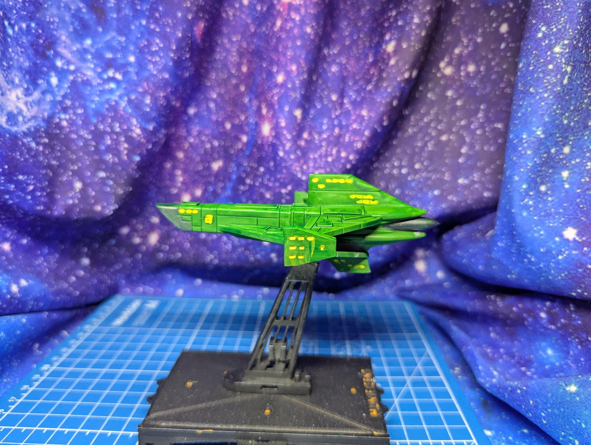 May include: A green spaceship model with yellow accents is displayed on a black stand. The spaceship has a sleek, angular design with wings and a pointed front. The model is painted in shades of green with yellow details. The stand is black and has a grid-like base.