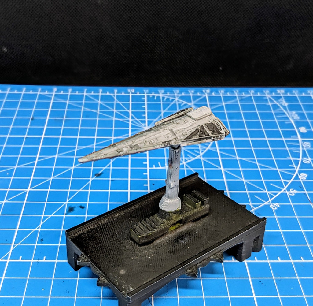 May include: A white and gray spaceship model on a black base. The spaceship is perched on a gray stand and has a pointed nose with a wing on each side. The base has a textured surface and is shaped like a platform with a step on one side.