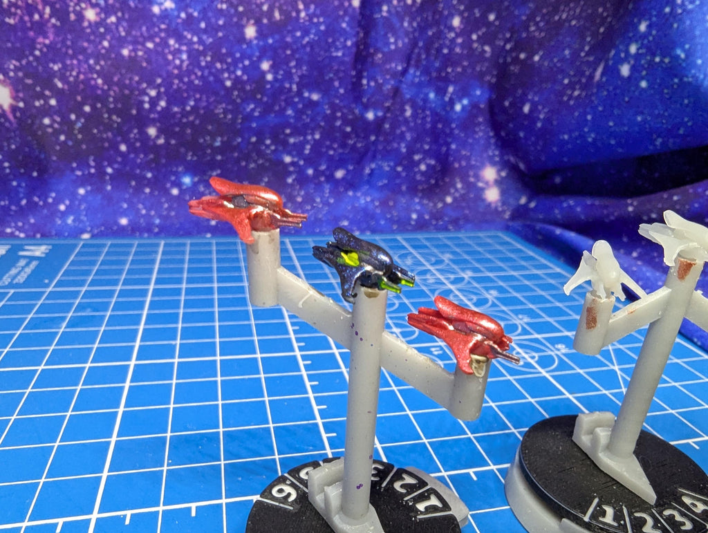 May include: Three miniature spaceships on display stands. One spaceship is red and black, one is green and black, and one is white. The stands are gray and have a circular base with numbers printed on them.