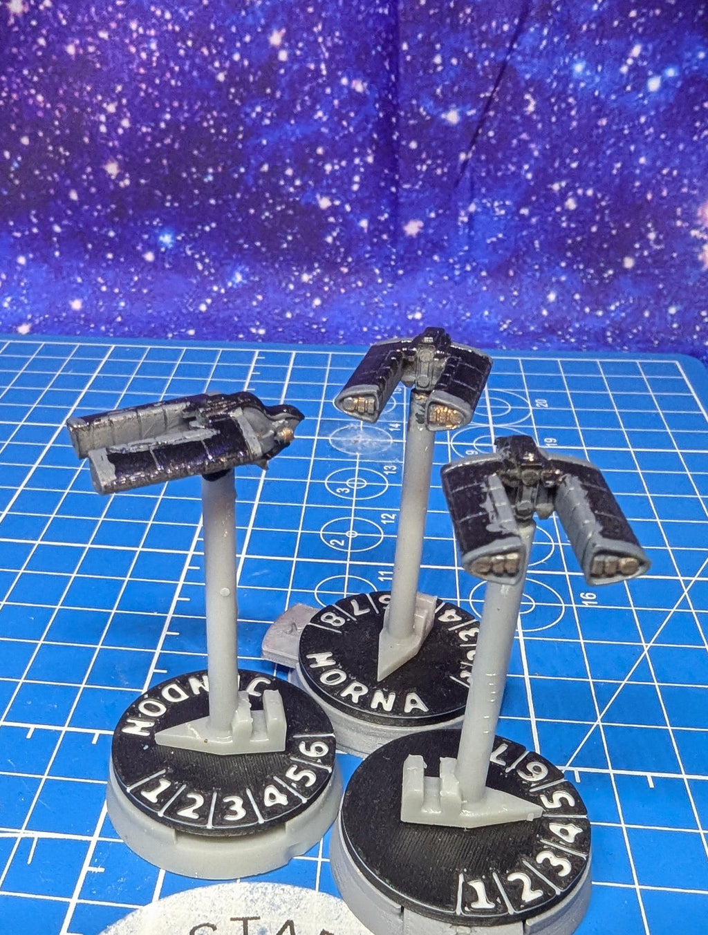 May include: Three miniature spacecraft models. Each model is black and gray, mounted on a gray stand attached to a circular base. The bases are black with white numbers and the word 'HORNA'. The background is a blue surface with a galaxy pattern.