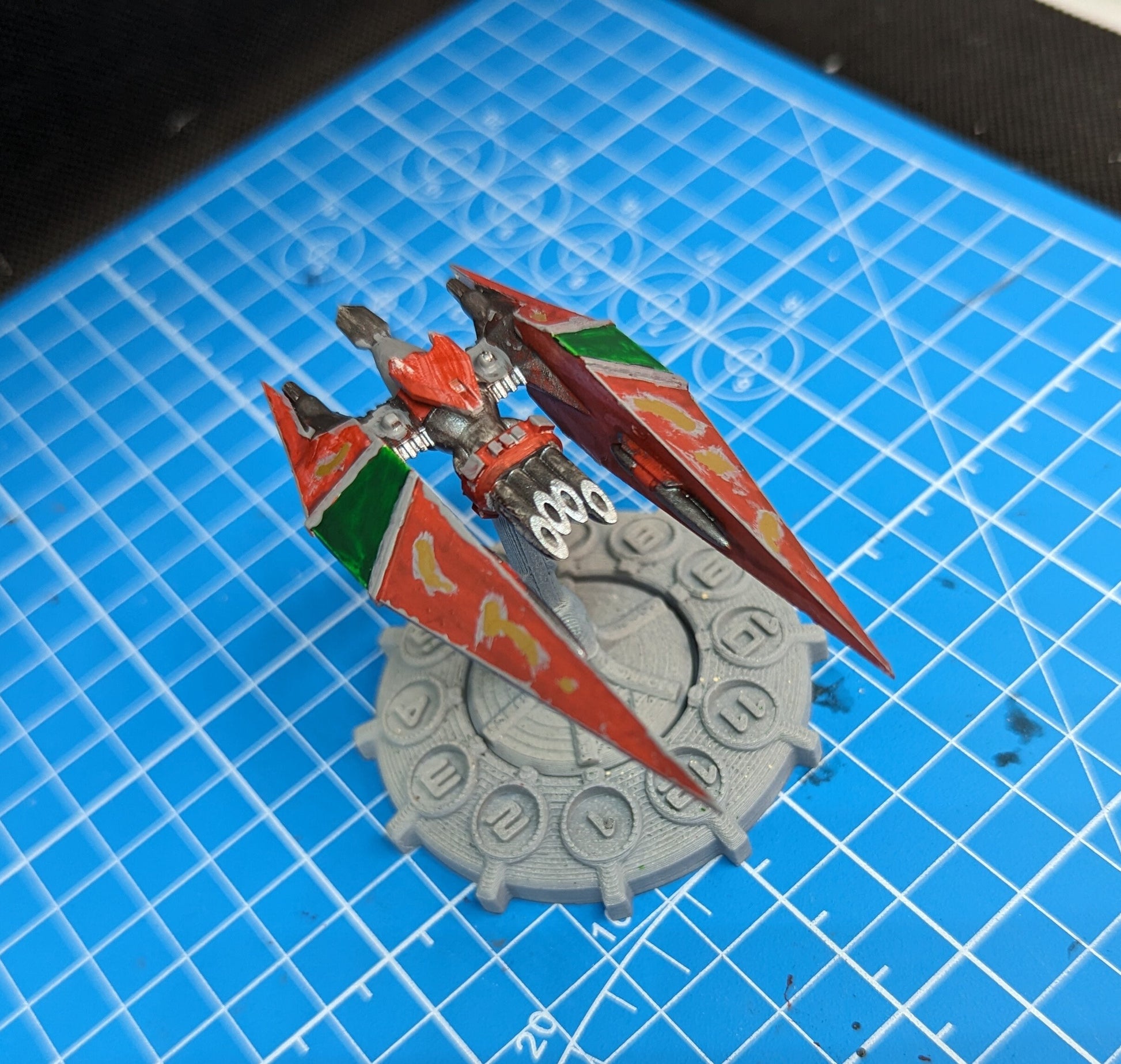 May include: A red, green, and gold spaceship model with a gray base. The spaceship has a pointed nose and two wings. The base has a circular design with numbers around the edge.