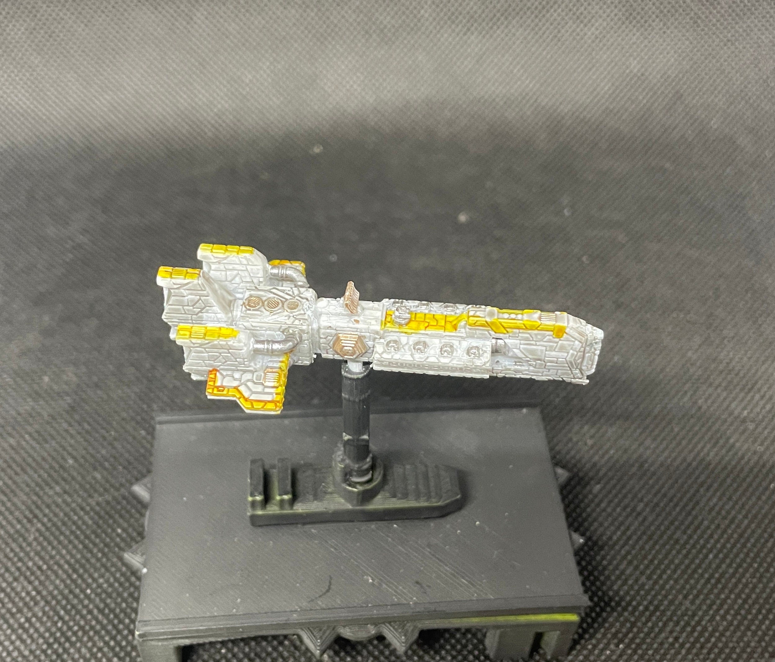 May include: A white and yellow spaceship model on a black base. The spaceship has a detailed design with a pointed nose and a long, thin body. The spaceship is painted in a silver and gold color scheme.